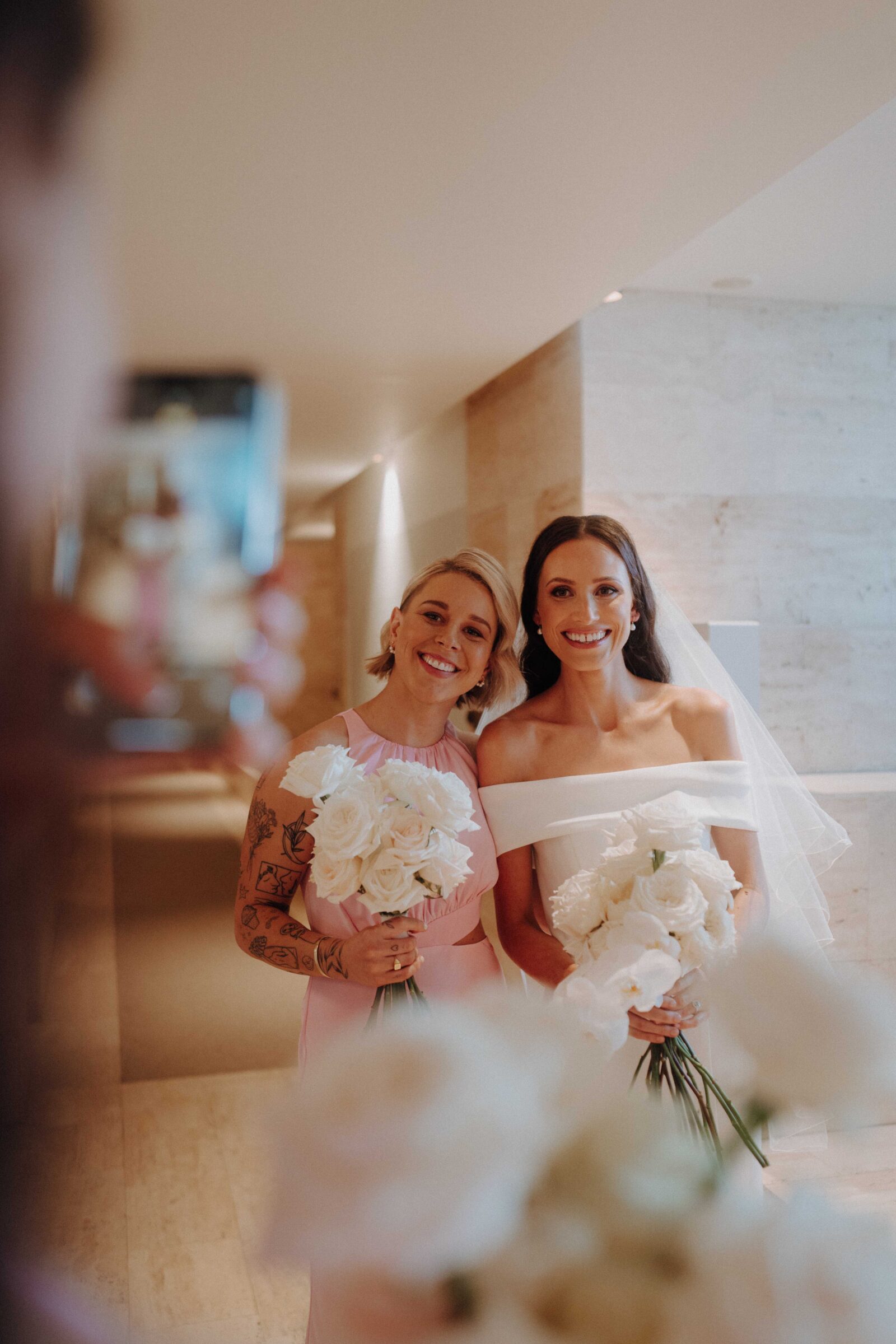 Two people in formal attire pose indoors with white roses, one wearing a white dress and veil, the other in a pink dress with tattoos, as a third person takes a photo with a phone.