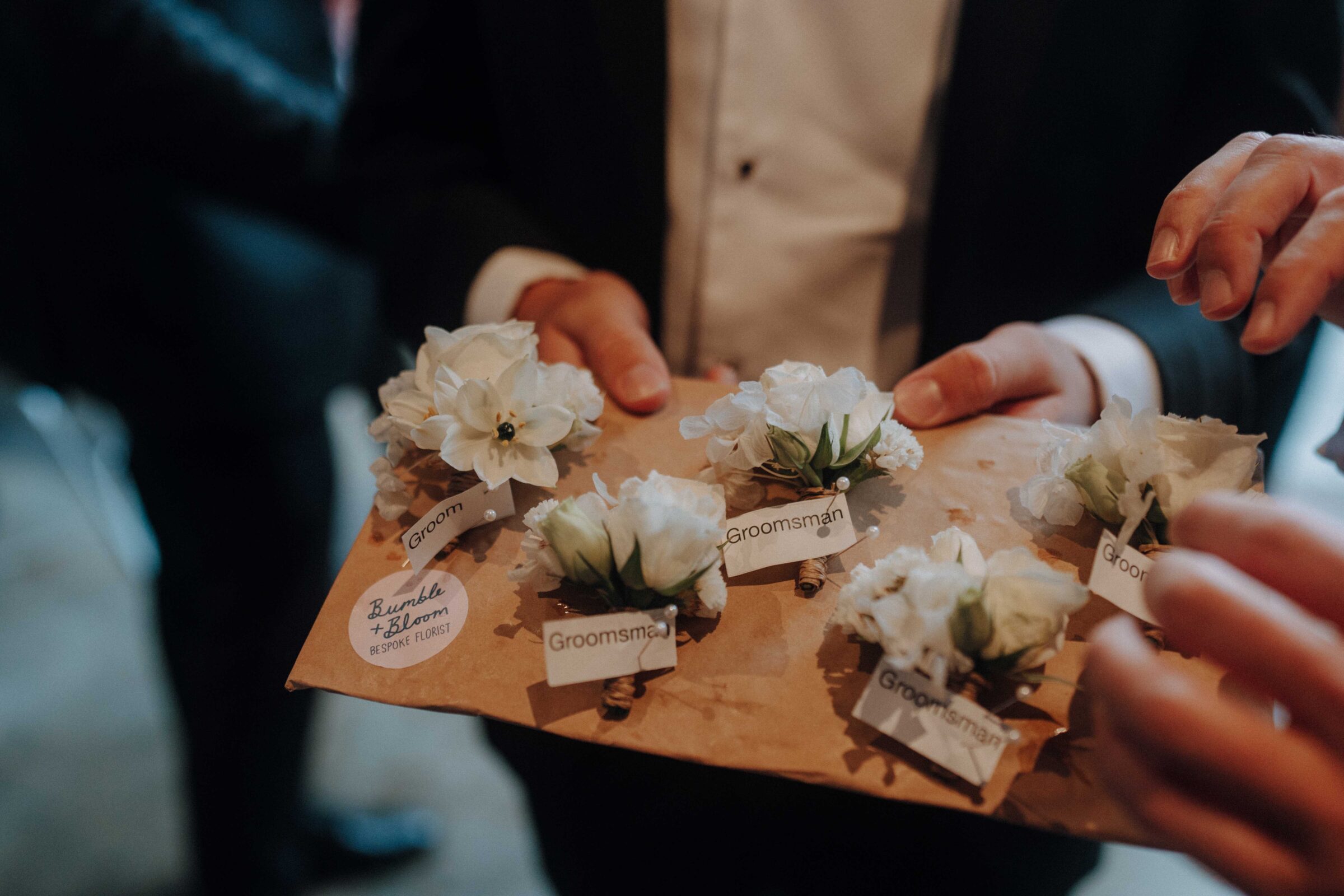 A person holds a tray with labeled boutonnieres for the groom and groomsmen, decorated with white flowers.