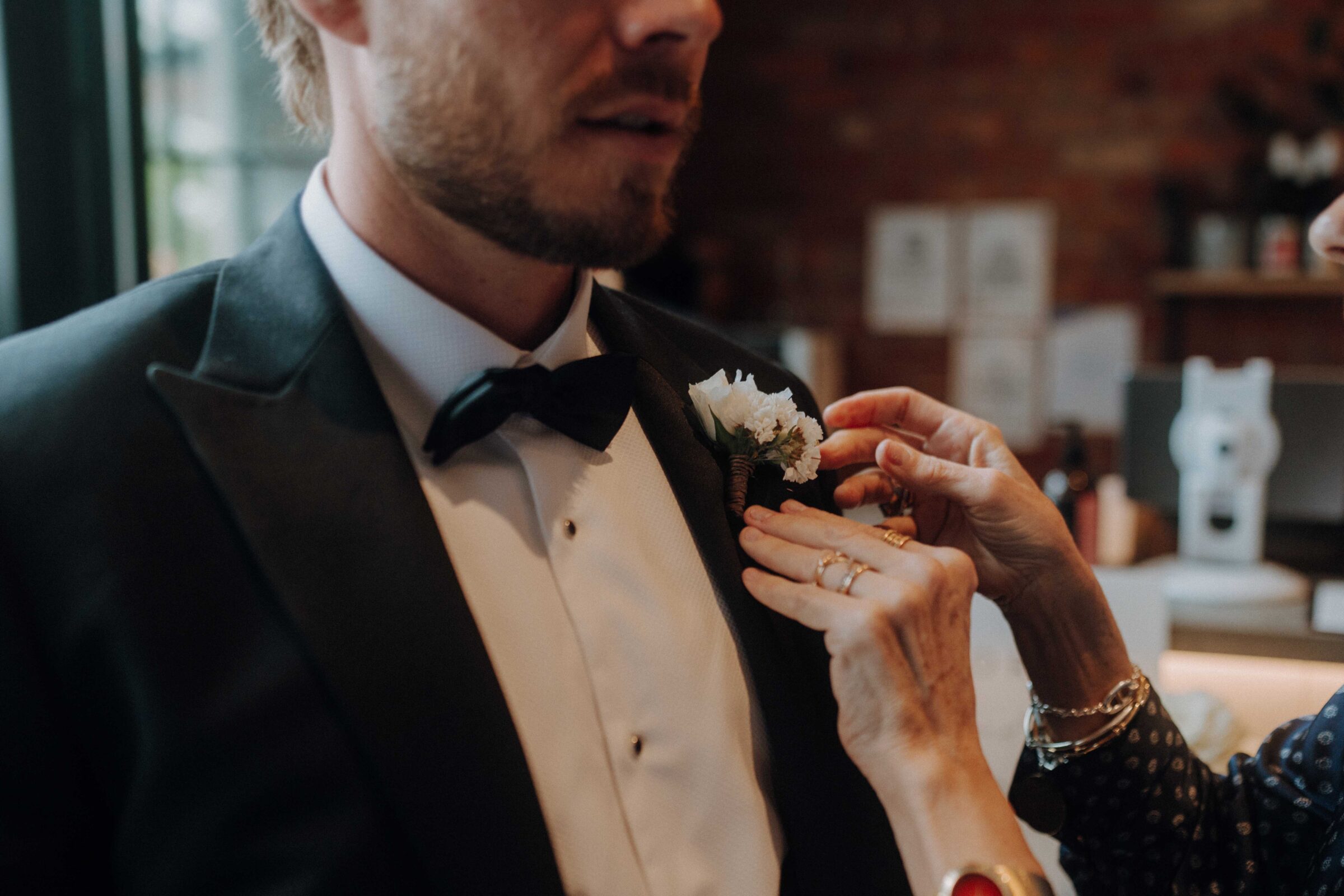 A person adjusts a boutonniere on a man wearing a tuxedo indoors.