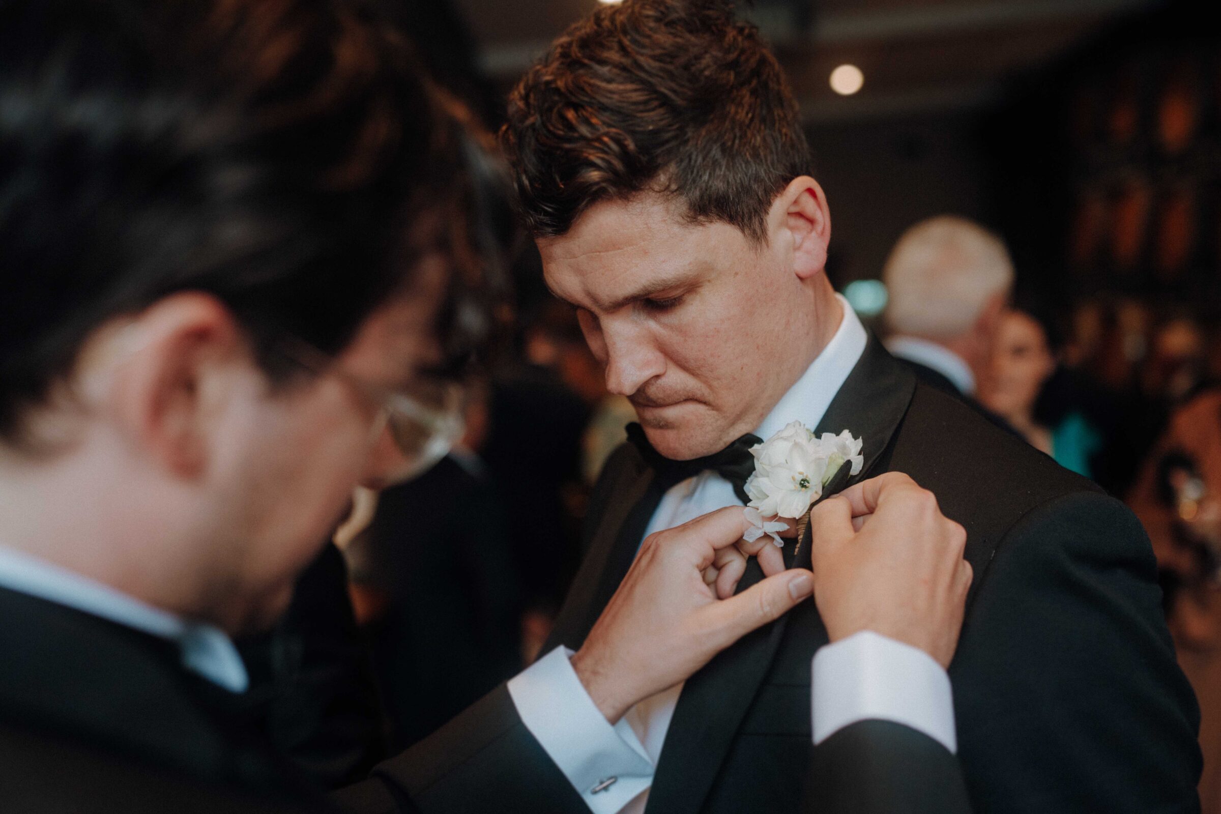 A man adjusts another man's boutonniere on a suit jacket at a formal event.