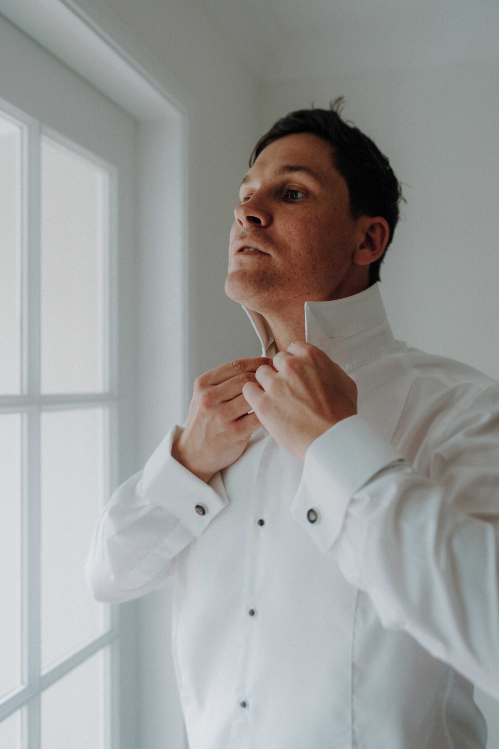 A person adjusts the collar of a white dress shirt while standing near a window.