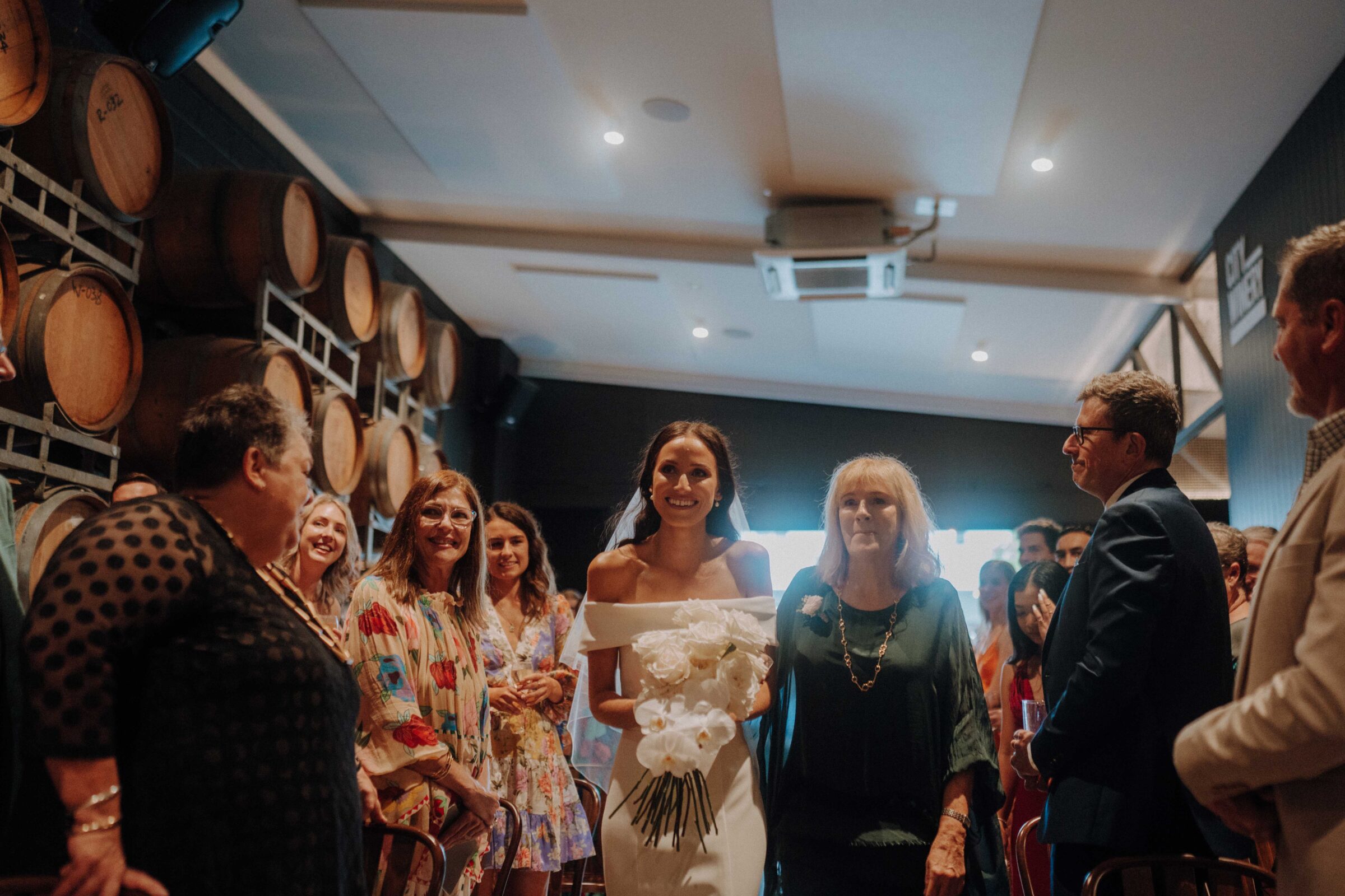 A bride in a white dress holds a bouquet, walking down the aisle flanked by guests in a wine cellar venue.