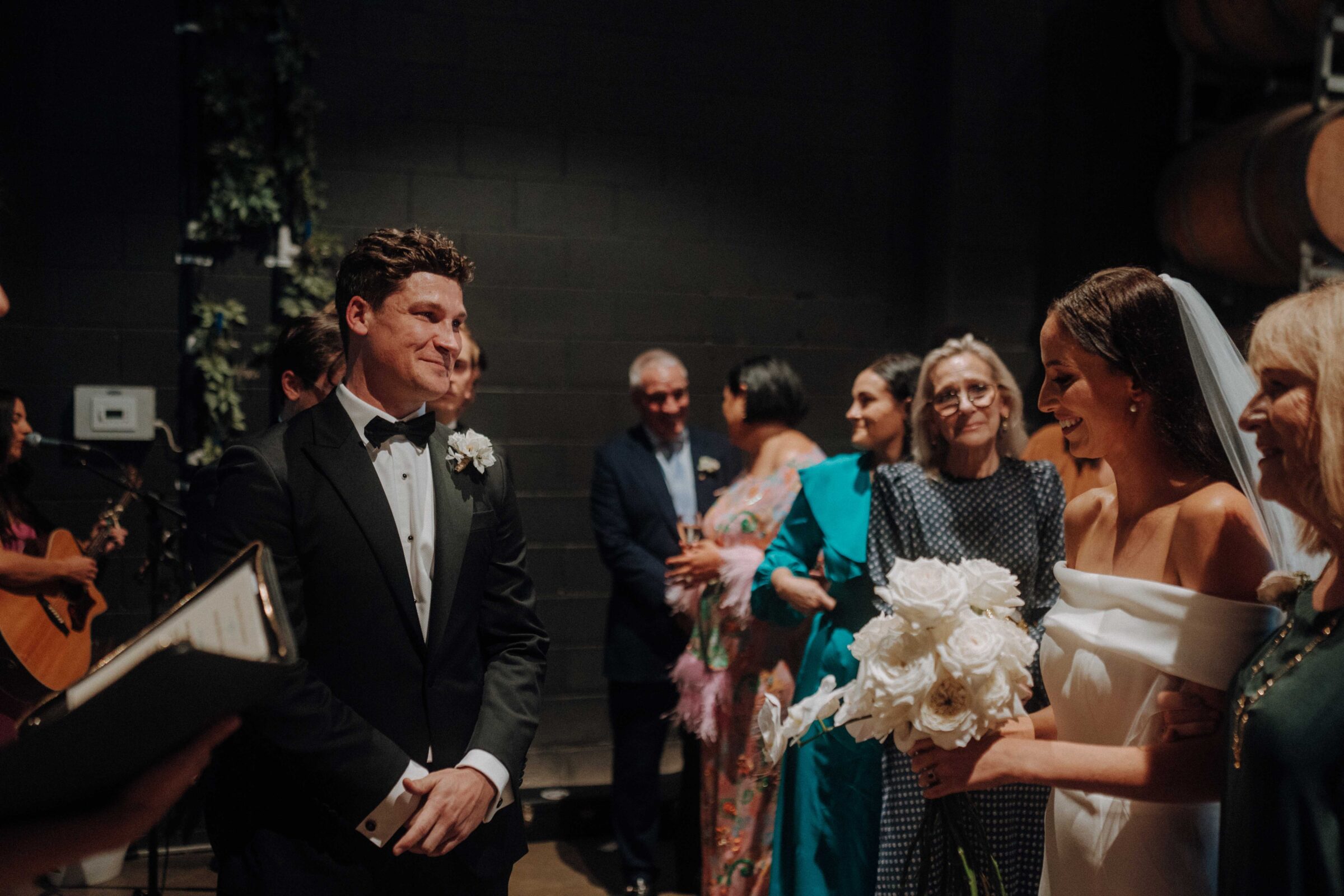 Bride and groom exchange smiles during their wedding ceremony, with guests looking on in the background.