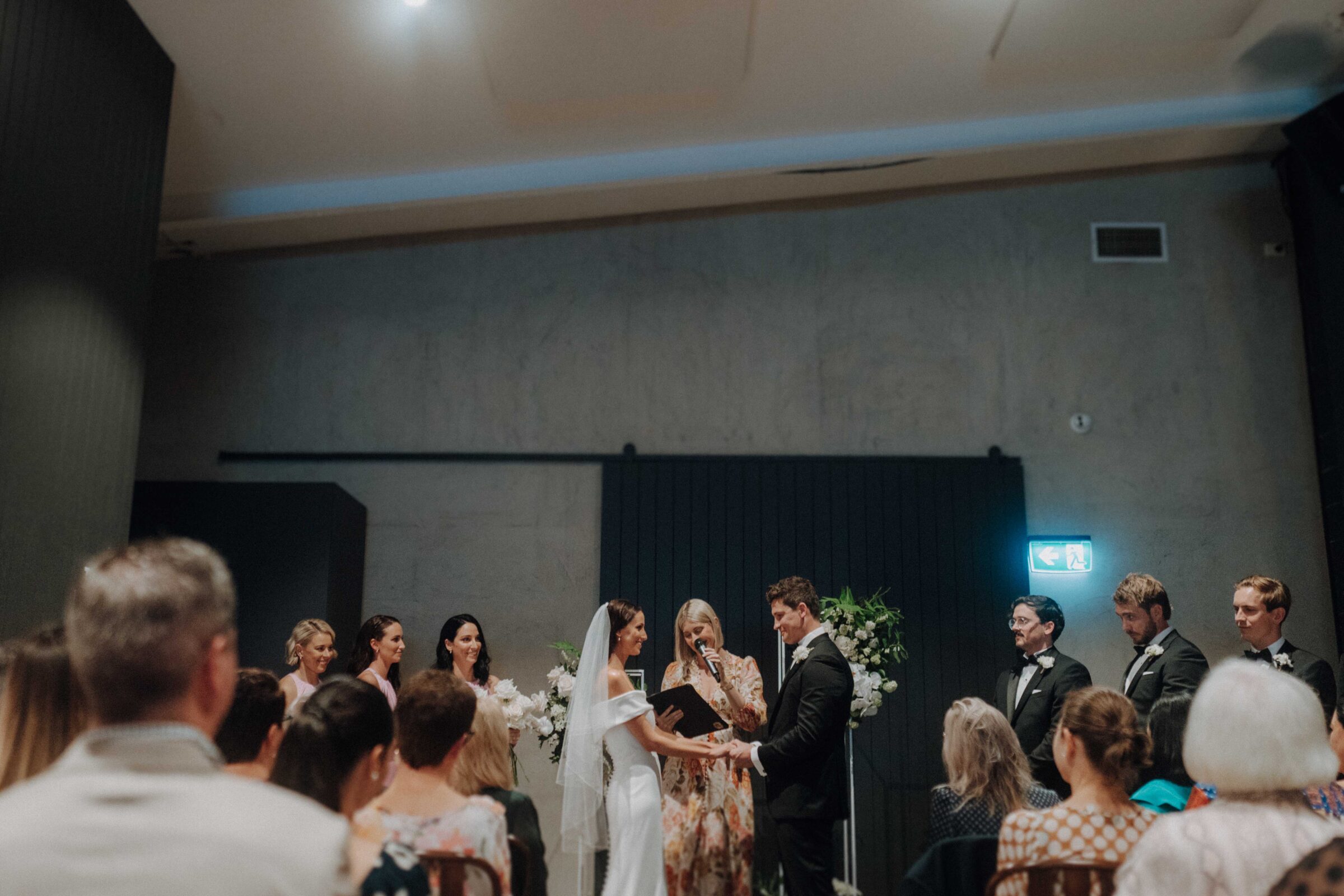 A wedding ceremony with a couple exchanging vows before an officiant. Bridesmaids and groomsmen stand on either side, with guests seated in the foreground.