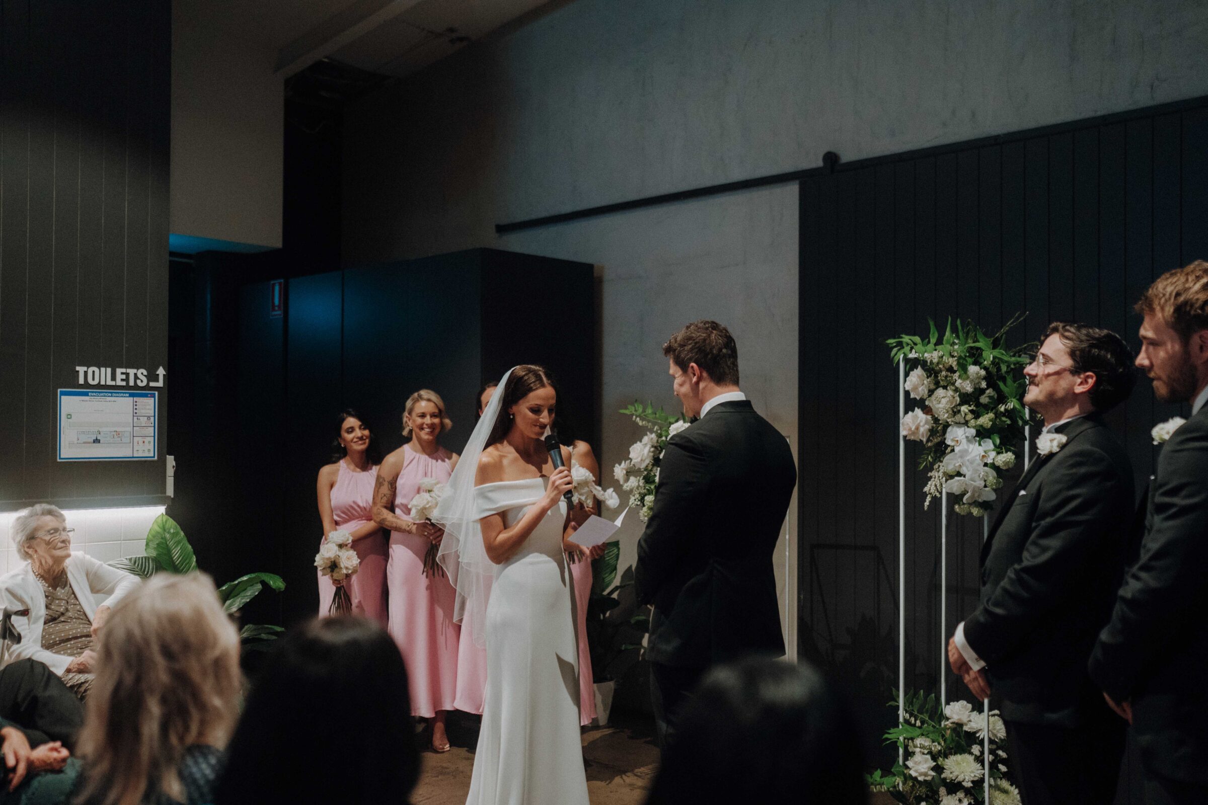 A bride and groom stand before each other in a wedding ceremony, with bridesmaids in pink dresses and groomsmen in black suits nearby.