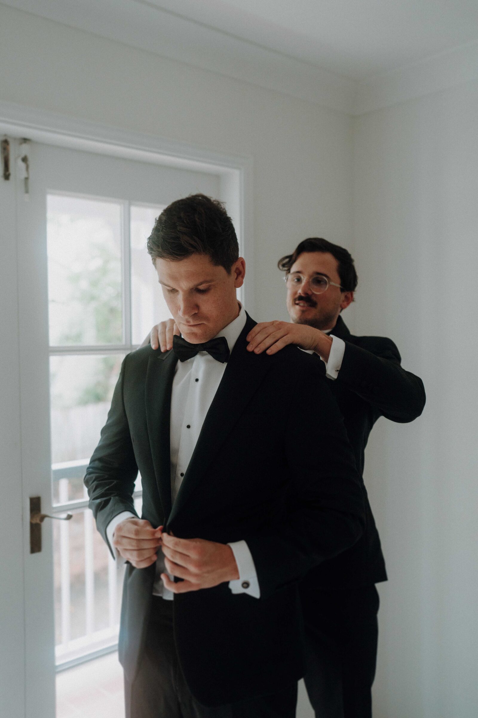 A man adjusts his bow tie while another man helps him with his suit jacket in a room with a door and window. Both are wearing formal attire.