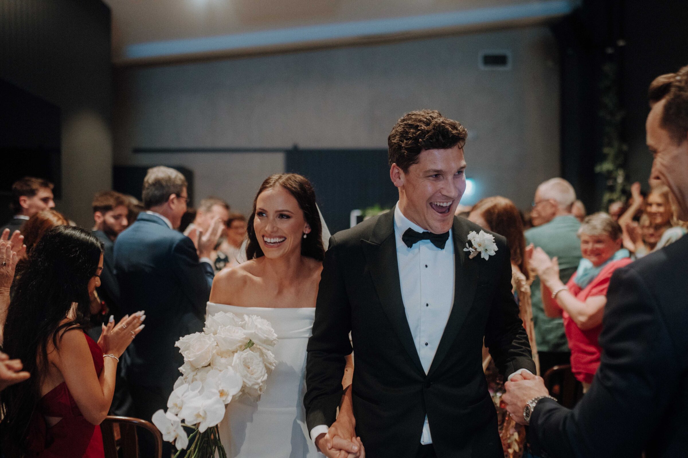 A bride and groom walk hand-in-hand down an aisle, smiling, with guests clapping on either side.