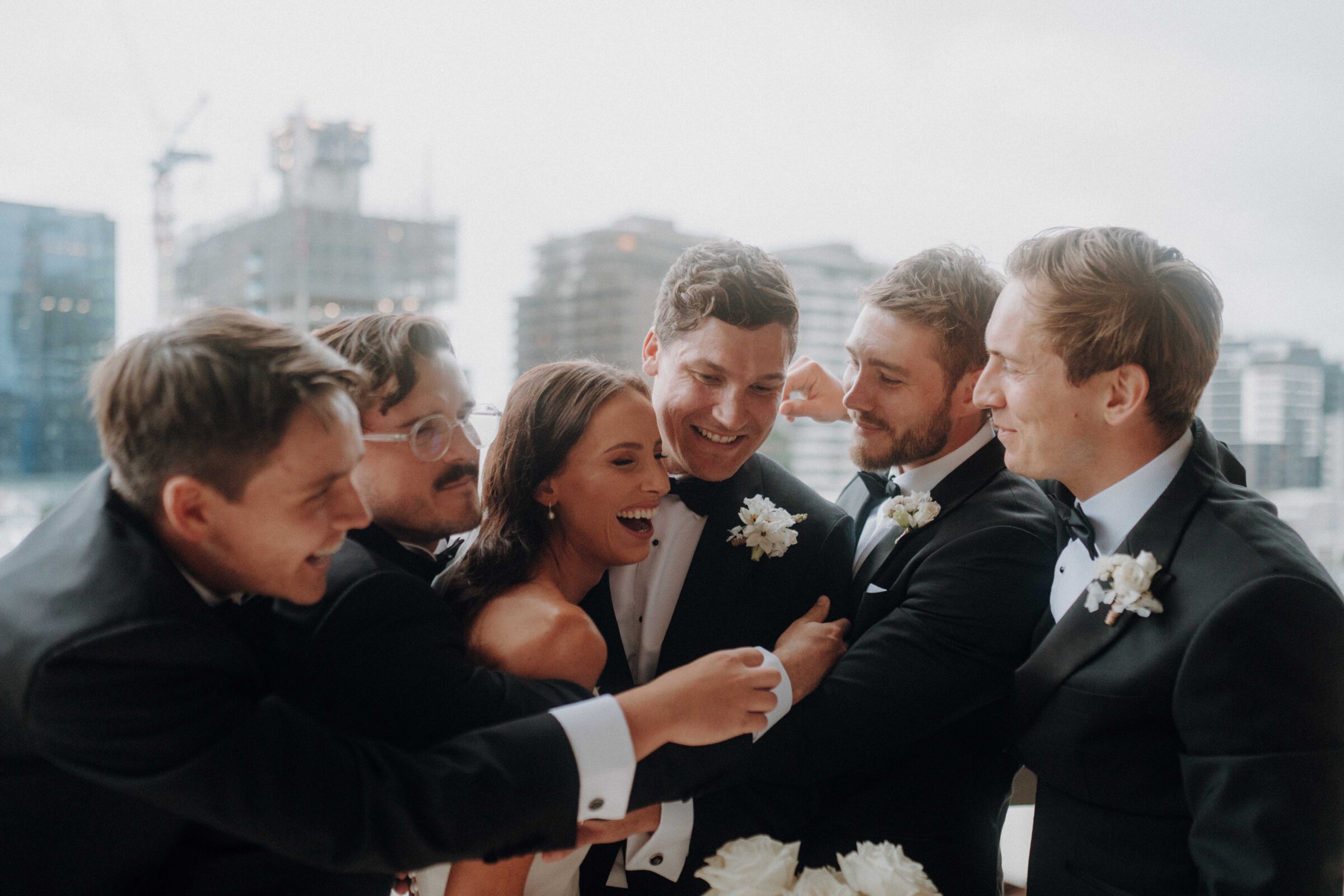 A bride and five groomsmen in tuxedos share a joyful group hug on a balcony with a cityscape background.