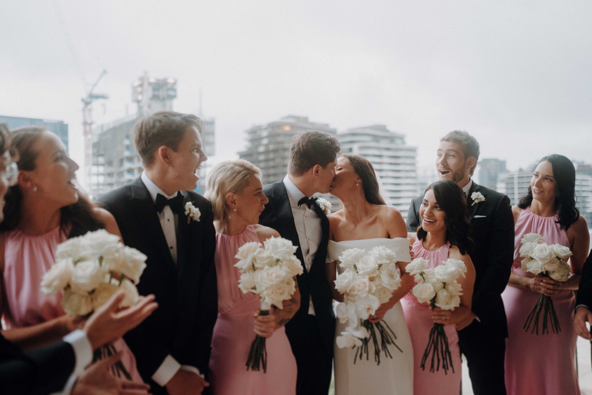 A wedding party with the couple kissing in the center, surrounded by bridesmaids in pink dresses and groomsmen in black suits holding white bouquets against a cityscape background.