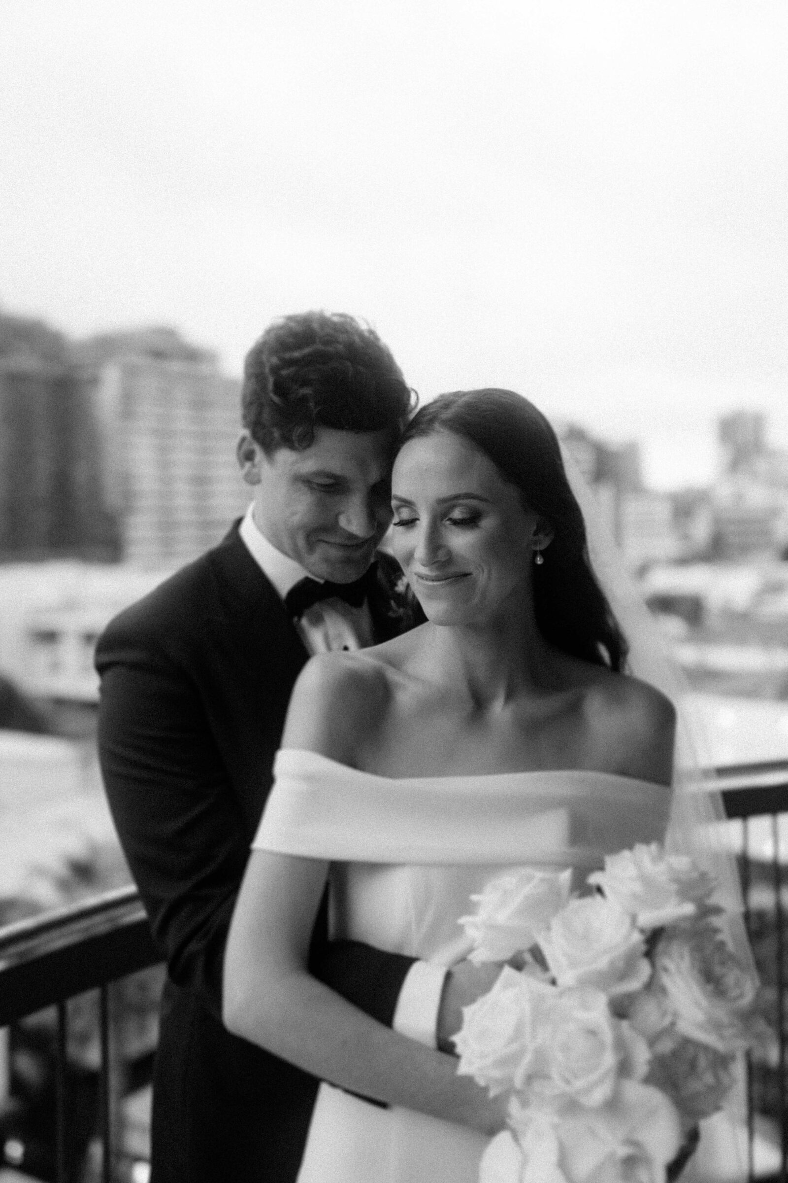 A bride and groom embrace on a balcony, with the groom standing behind the bride holding her bouquet. Both are smiling softly. The background shows an urban landscape.