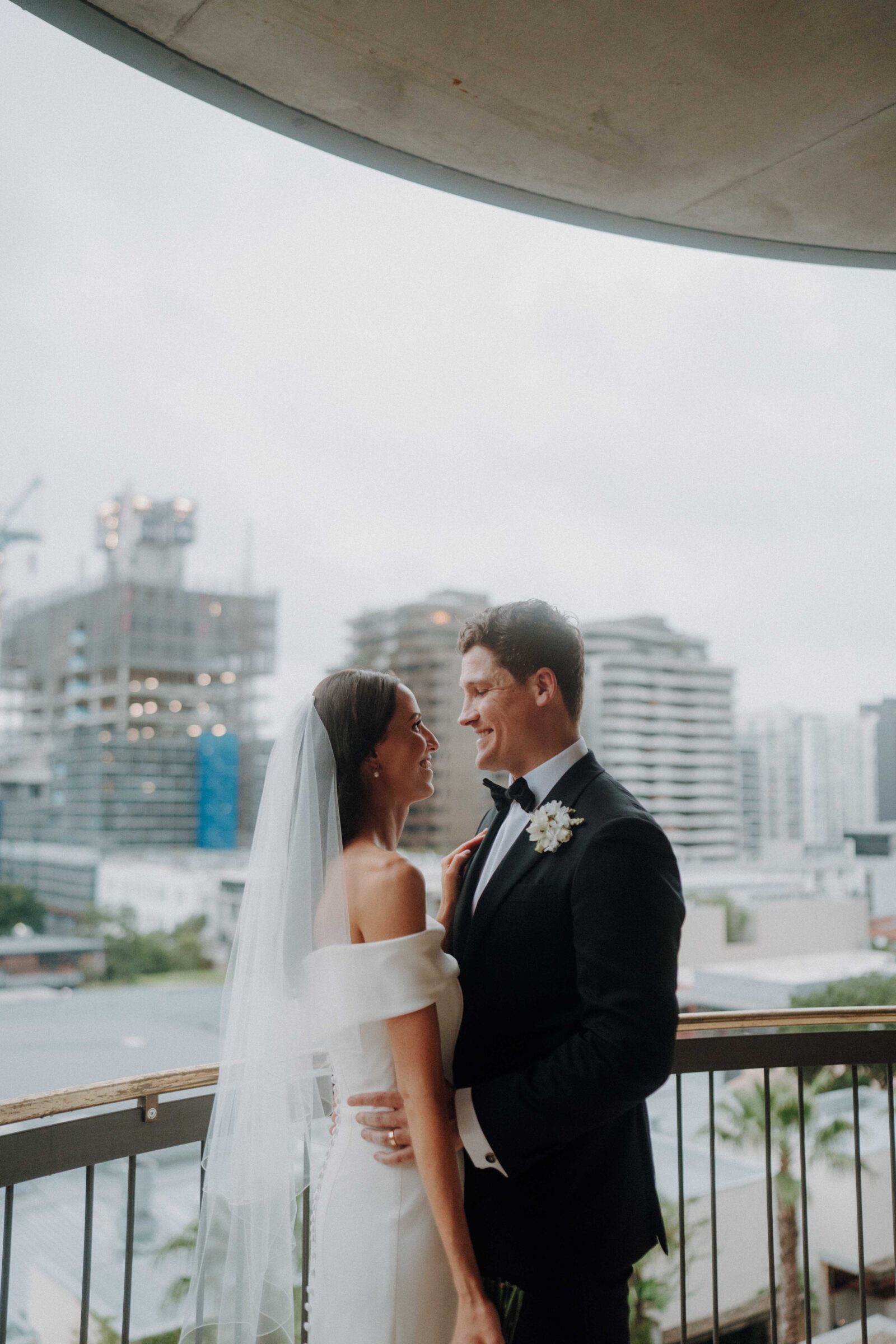 A bride and groom stand on a balcony, smiling at each other, with a cityscape and construction in the background.