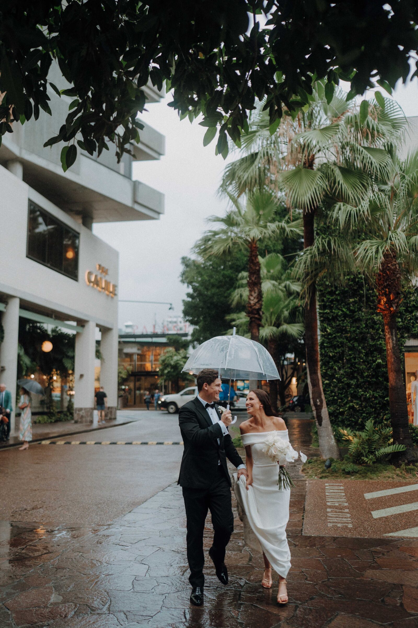 A couple walks in the rain sharing a transparent umbrella, with modern buildings and palm trees in the background.