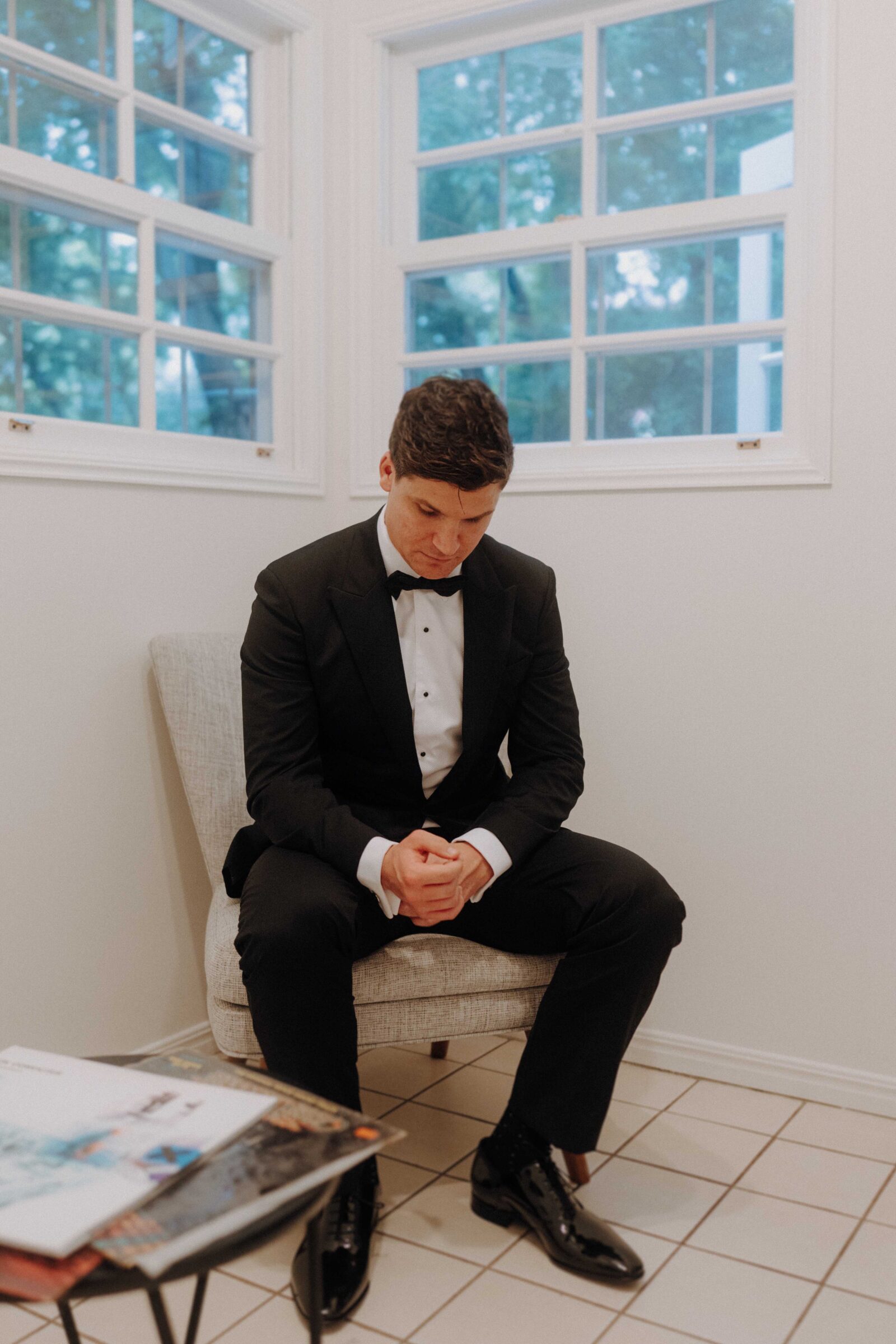 A man in a formal black suit and bow tie sits on a chair in a room with tiled floor and large windows, looking down with hands clasped. Nearby is a table with magazines.