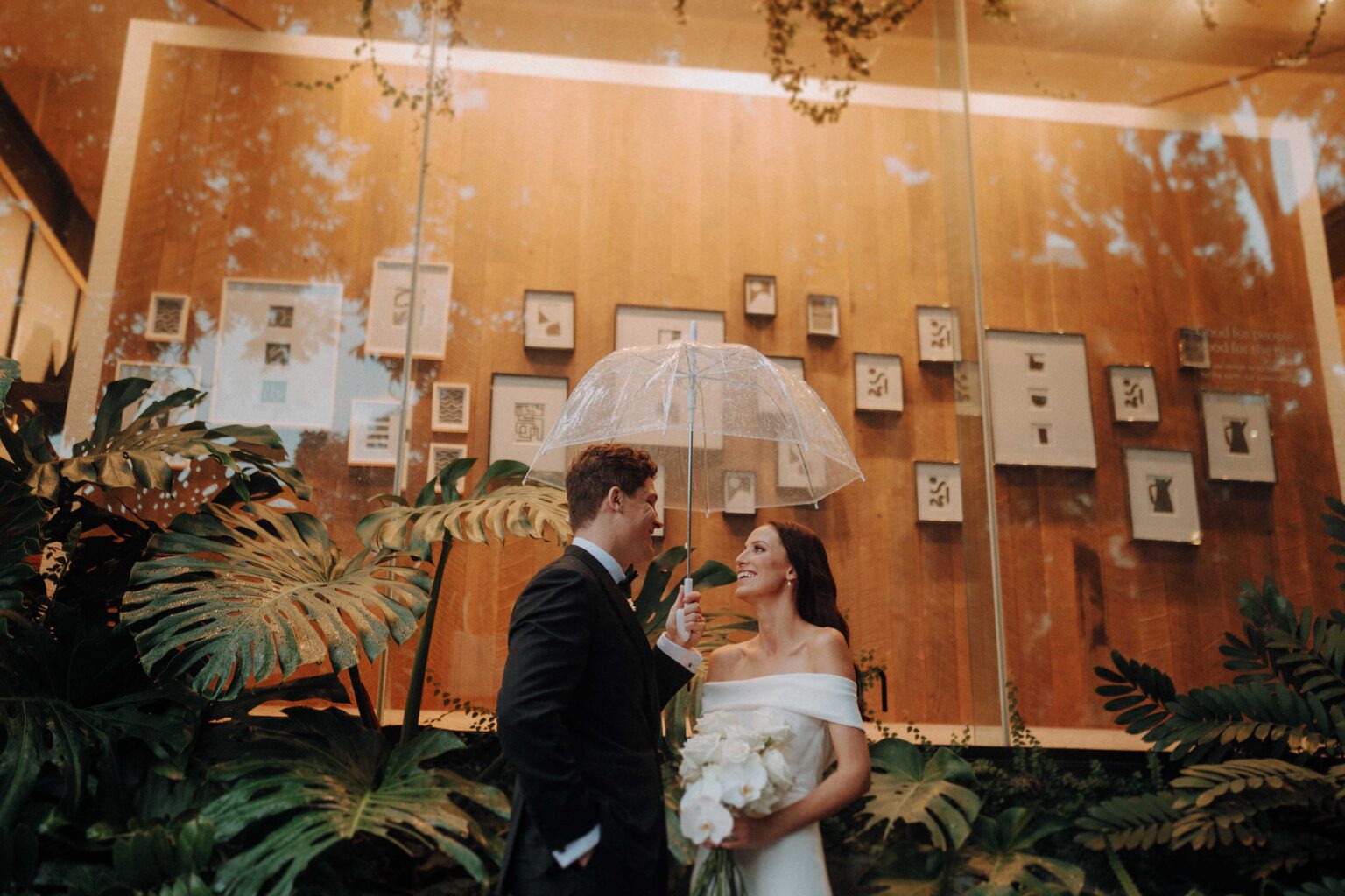 A couple stands under a clear umbrella, surrounded by lush greenery and framed artwork on a wooden wall.