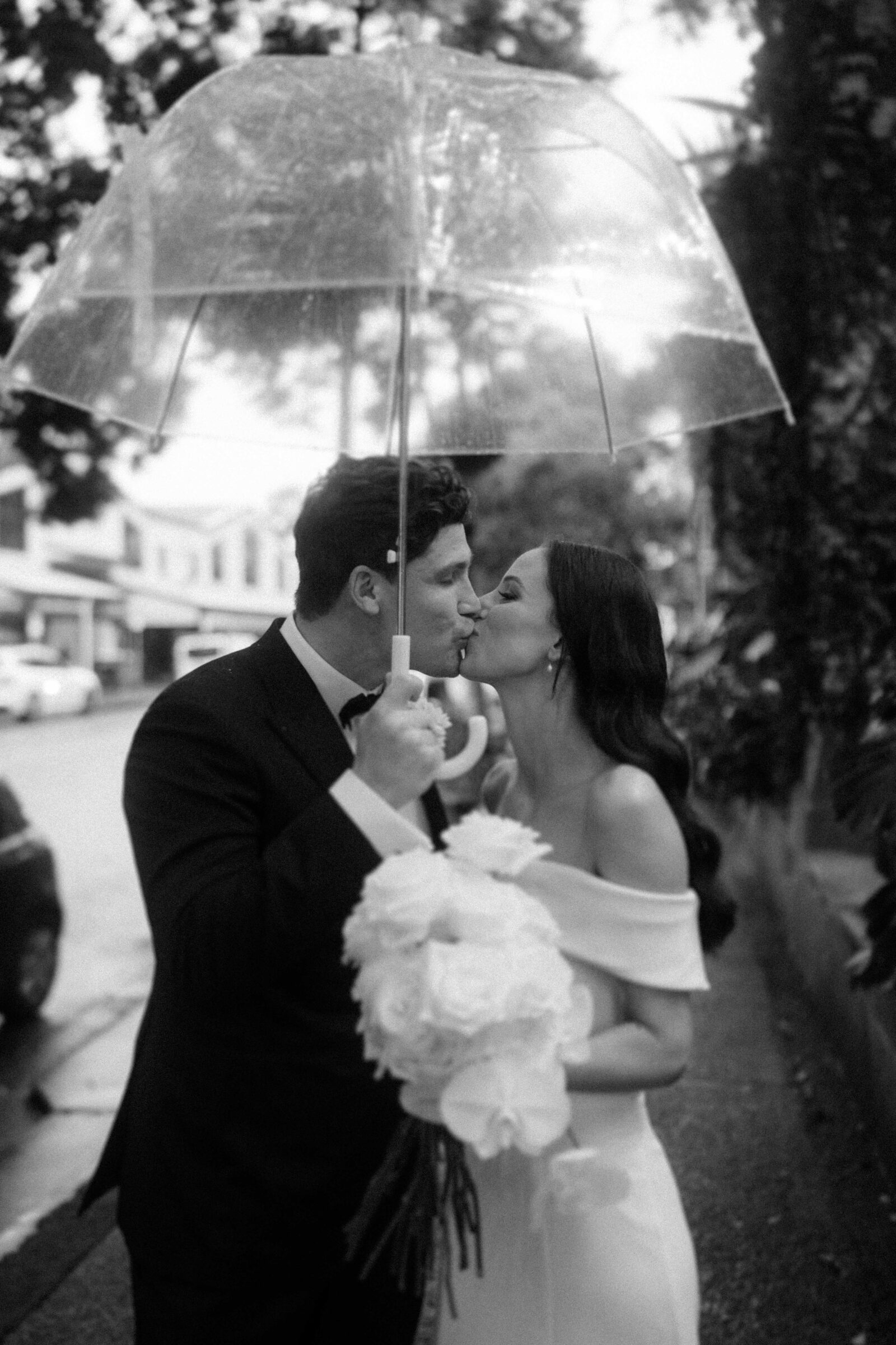 A couple, dressed in wedding attire, kisses under a clear umbrella. The bride holds a bouquet of white flowers. The background shows a street scene with trees and buildings.