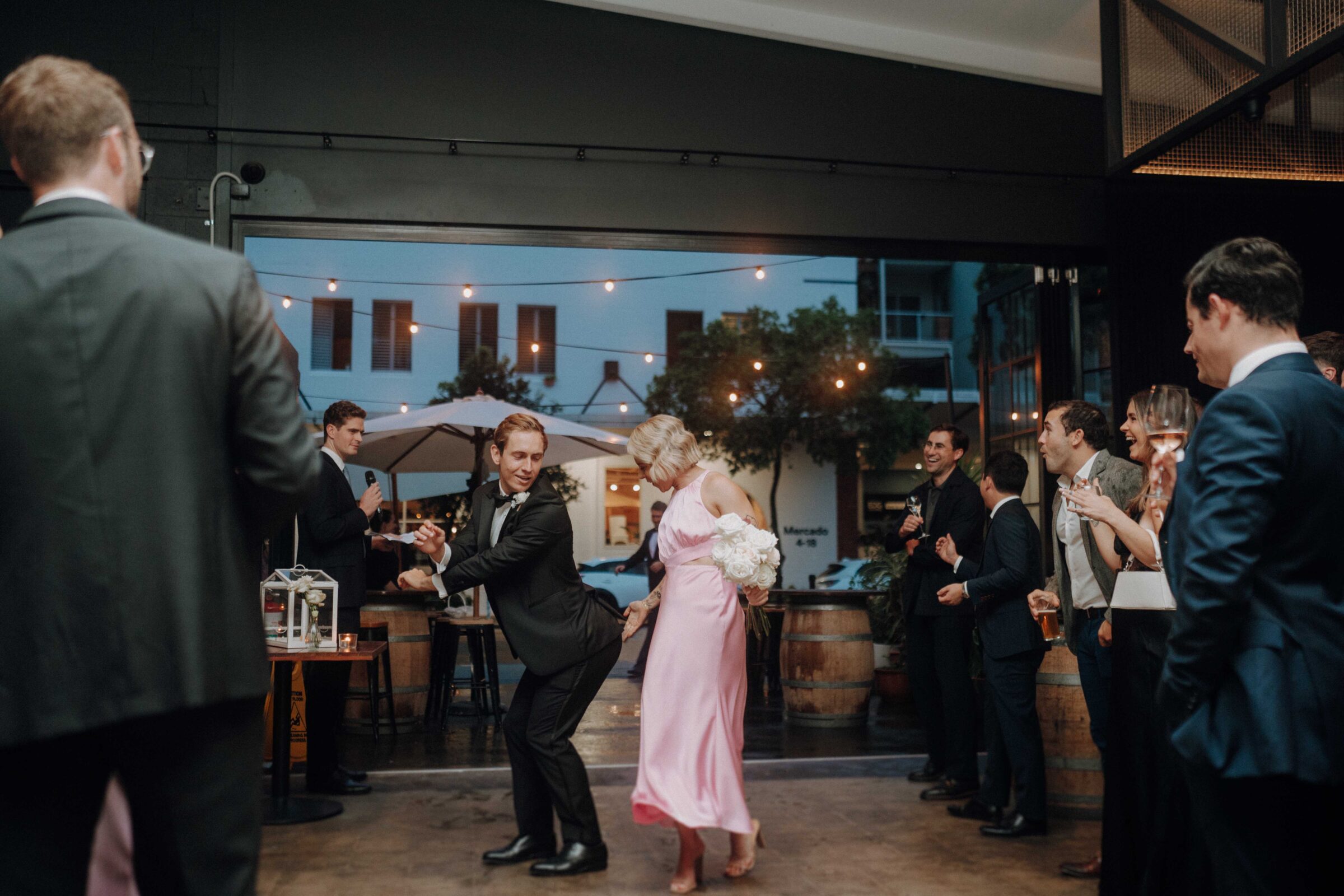 A couple dances enthusiastically in a room filled with people in formal attire. Others watch and smile. A band plays in the background.