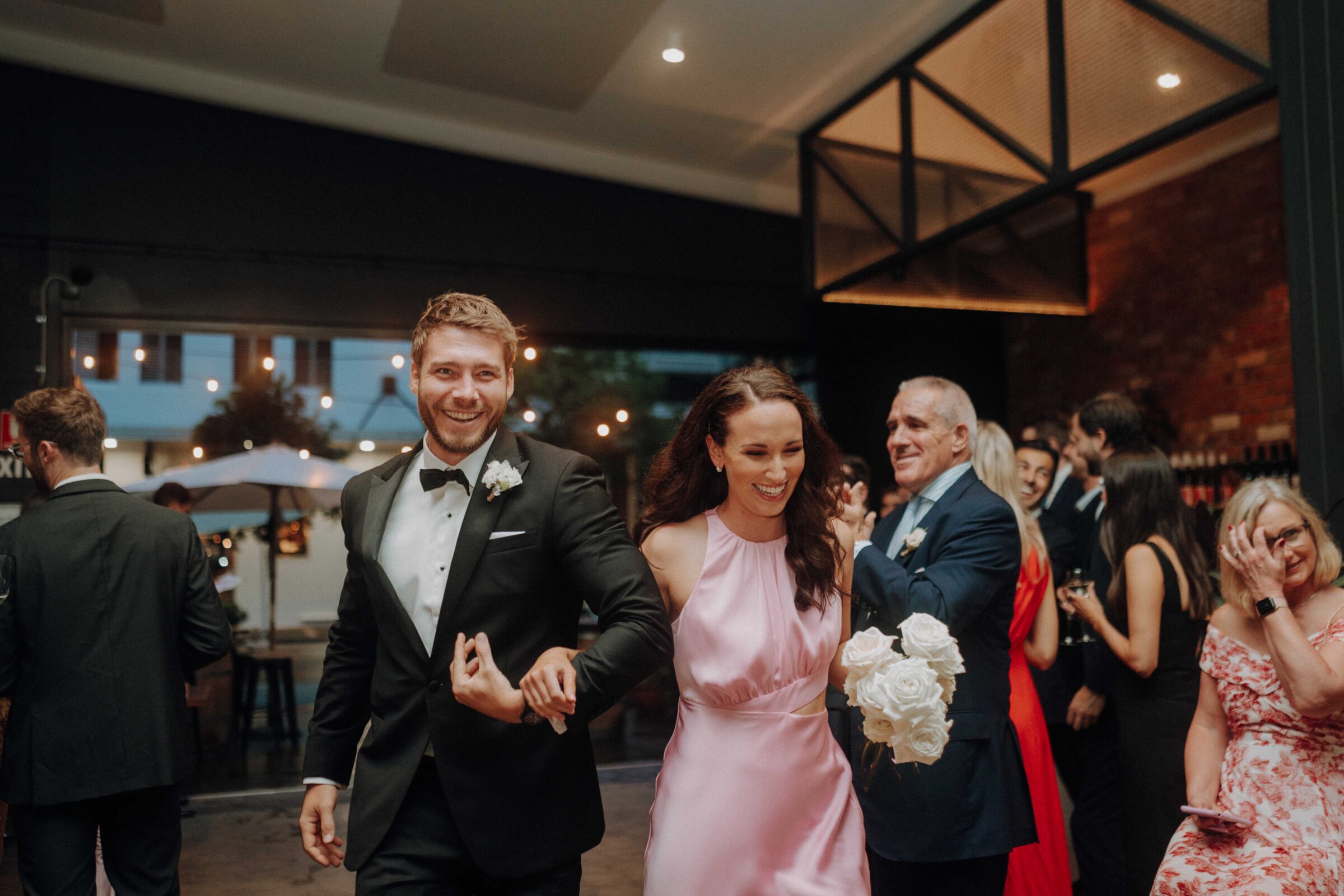 A smiling couple in formal attire enters a reception hall. The man wears a tuxedo, and the woman is in a pink dress, holding white flowers. Guests in the background appear joyful.