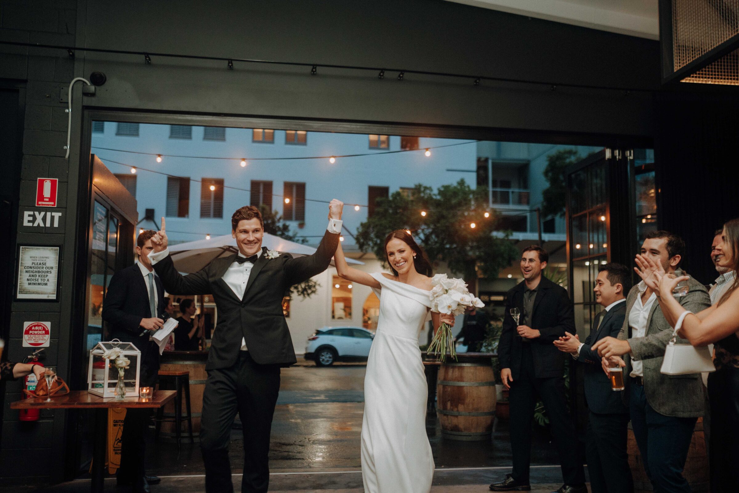 A bride and groom in formal attire hold hands and smile while entering a venue, with guests applauding around them.