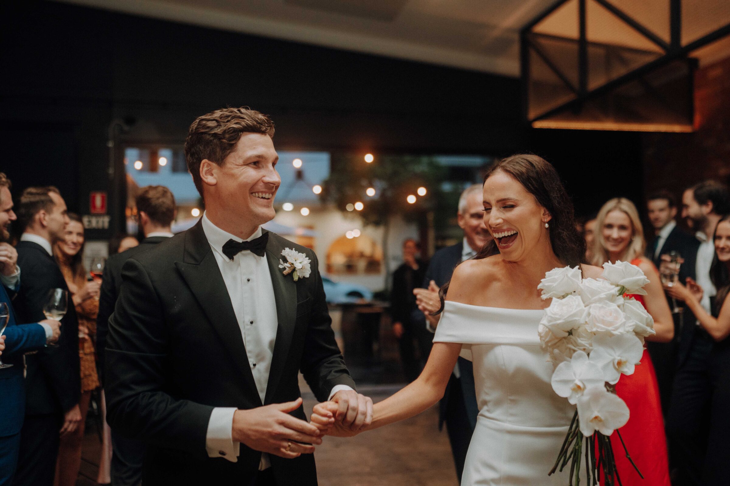 A bride and groom smiling and holding hands, walking through a crowd of well-dressed guests. The bride holds a bouquet of white flowers.