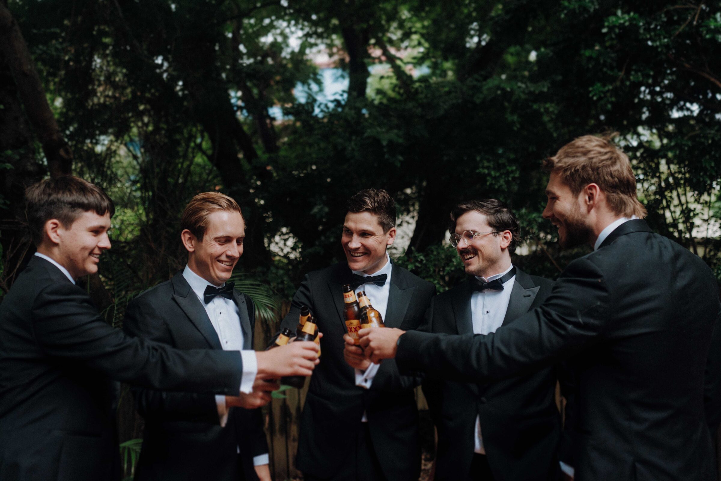 Five men in suits clink beer bottles together outdoors, surrounded by trees, while smiling at each other.