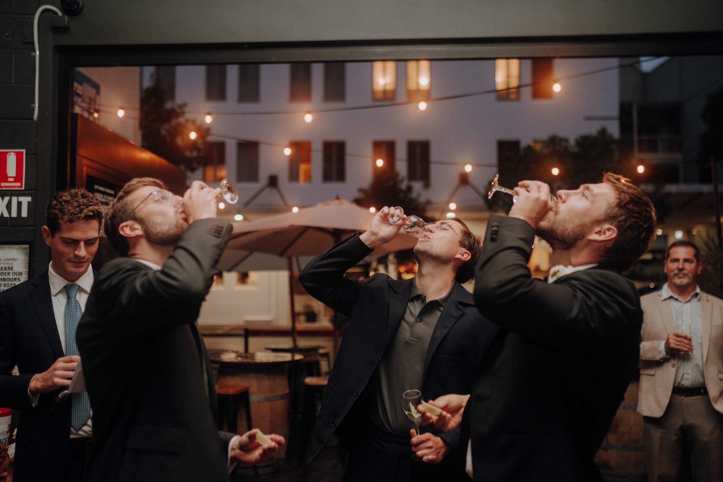 A group of five men in suits standing indoors; four are drinking from glasses while one watches.