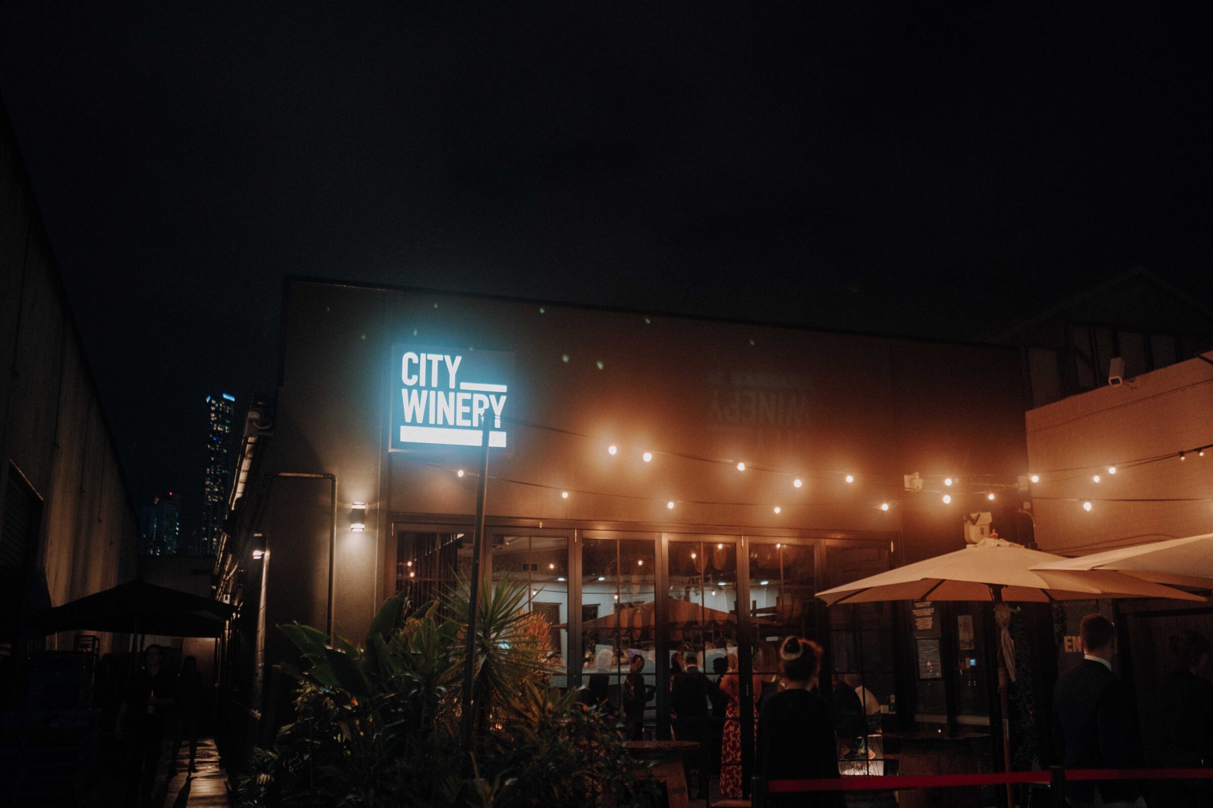 Outdoor view of City Winery at night, featuring string lights, plants, umbrellas, and people gathered outside.