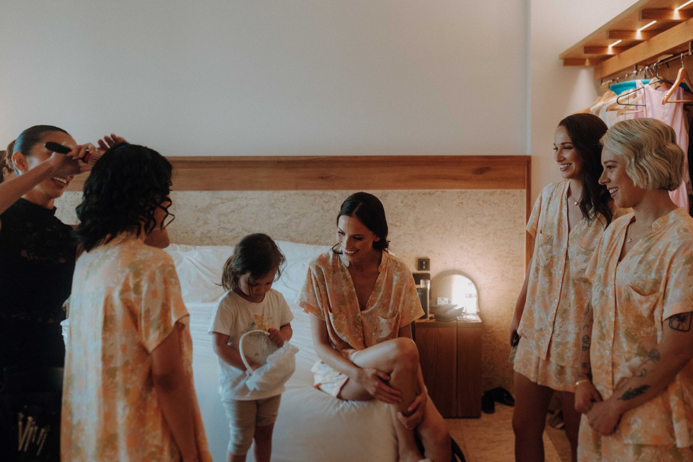 A group of women and a child in matching robes gather around a bed in a warmly lit room.