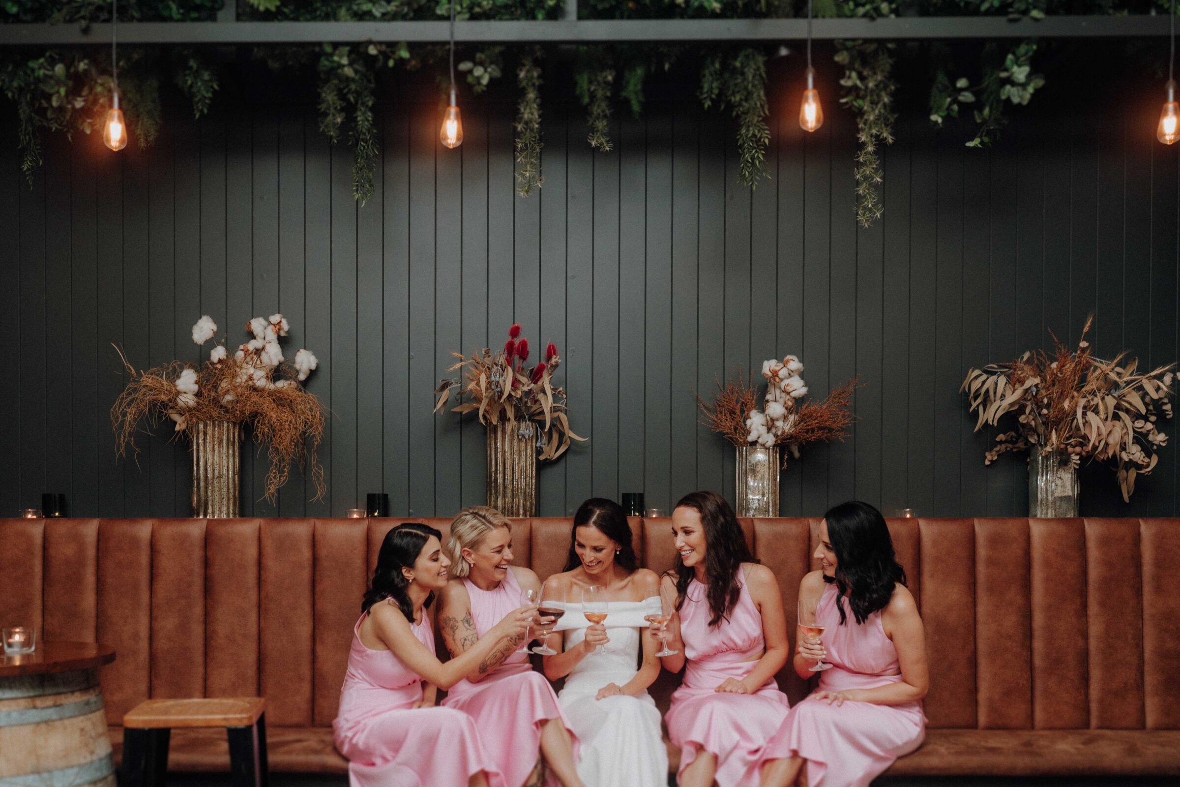 A bride in a white dress sits on a leather couch with four bridesmaids in pink dresses, holding drinks. The background features hanging lights and decorative vases with plants.