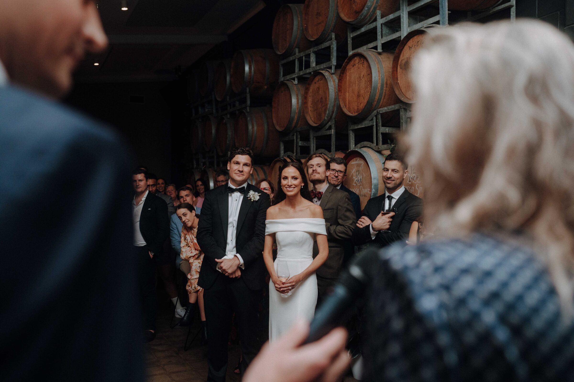 A couple stands facing a woman with a microphone during a wedding ceremony in a room with wooden barrels. Guests are seated and standing in the background.