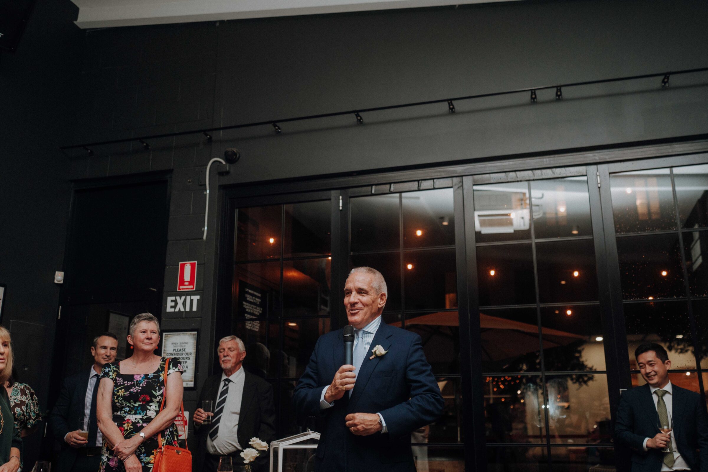 An older man in a suit speaks into a microphone at an indoor event. Several people stand nearby, listening.