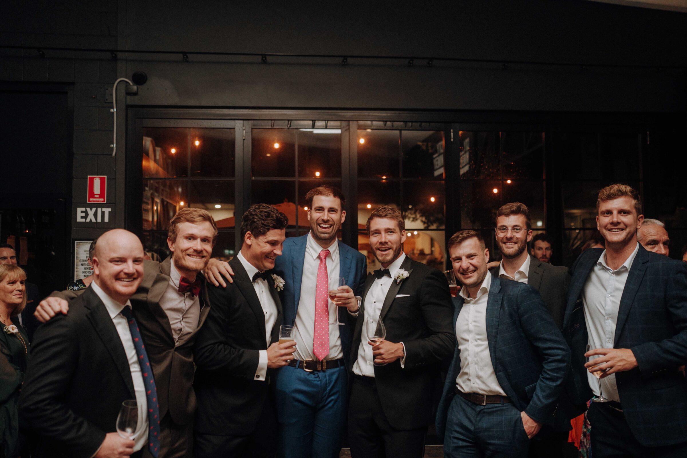 A group of eight men in suits posing together indoors, with dim lighting and a large window in the background. Some are holding drinks.