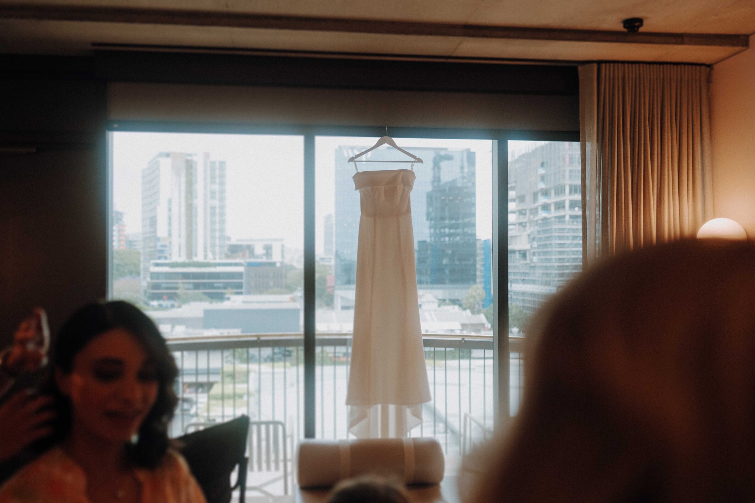 Wedding dress hanging in front of large windows with a cityscape view in the background.