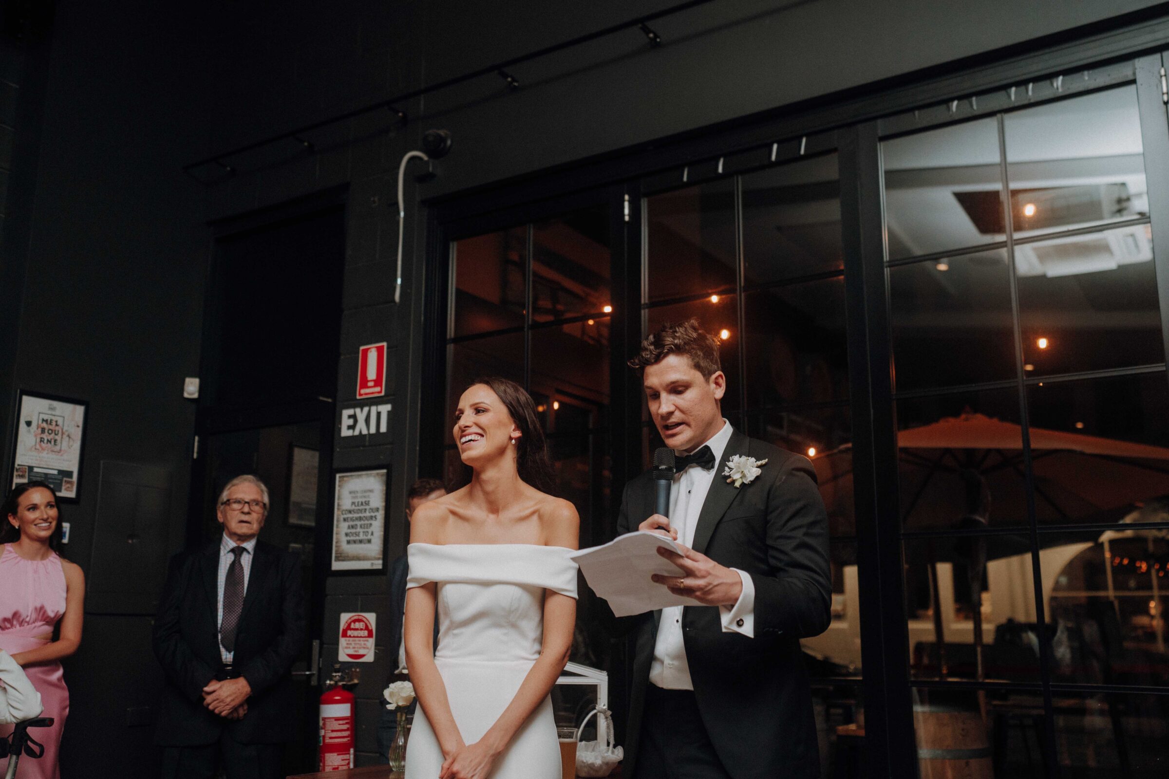 A bride and groom stand together indoors, with the groom holding a microphone and paper. They are dressed in wedding attire. A few people are visible in the background.