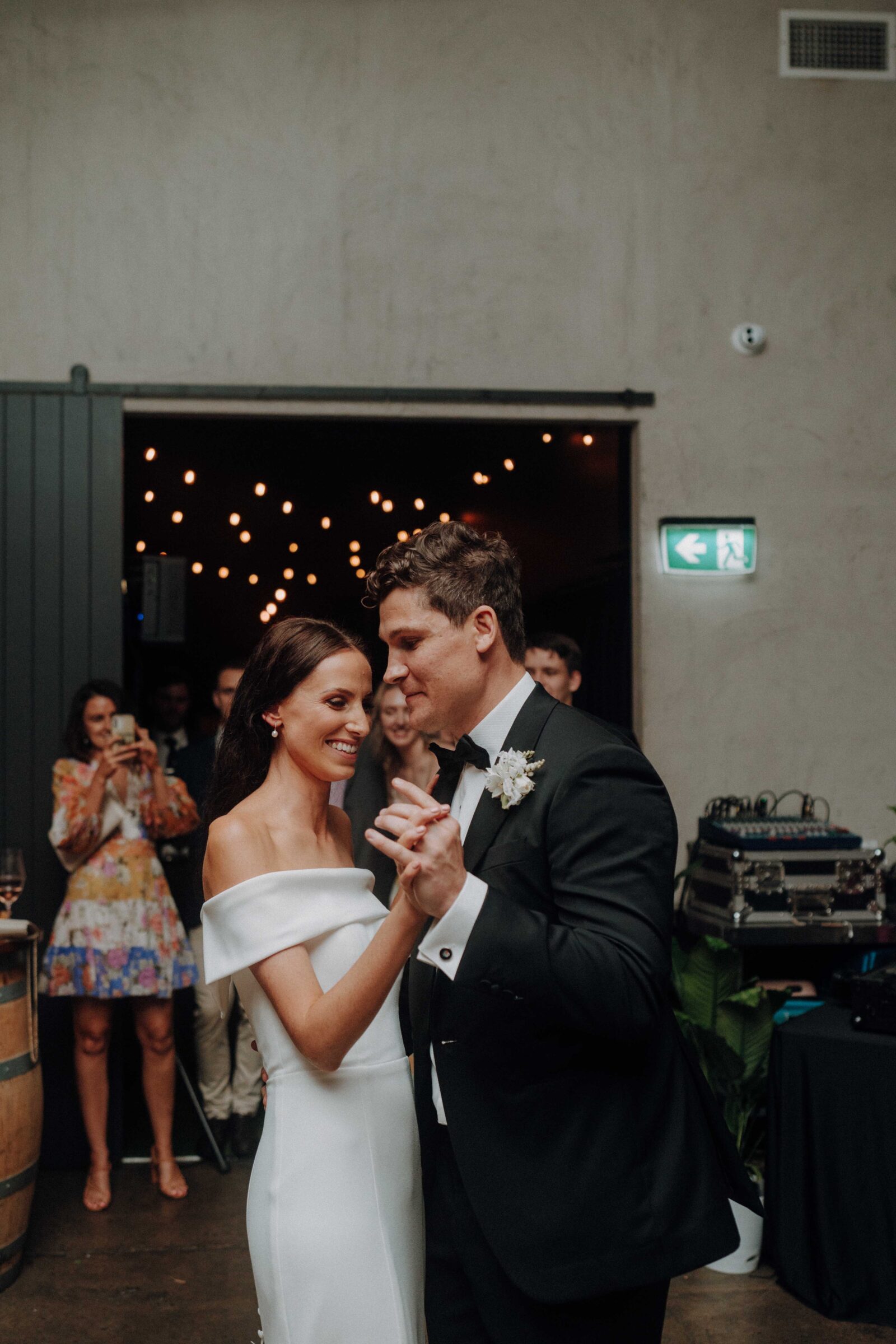 A couple dances together at a wedding, surrounded by guests. The bride wears a white dress, and the groom wears a black suit. There are decorative lights in the background.