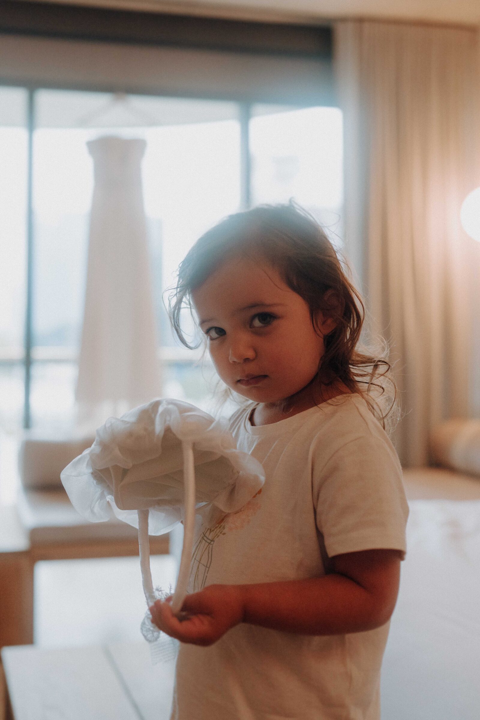 A young child in a t-shirt holds a white hat indoors, with a dress hanging in the background.