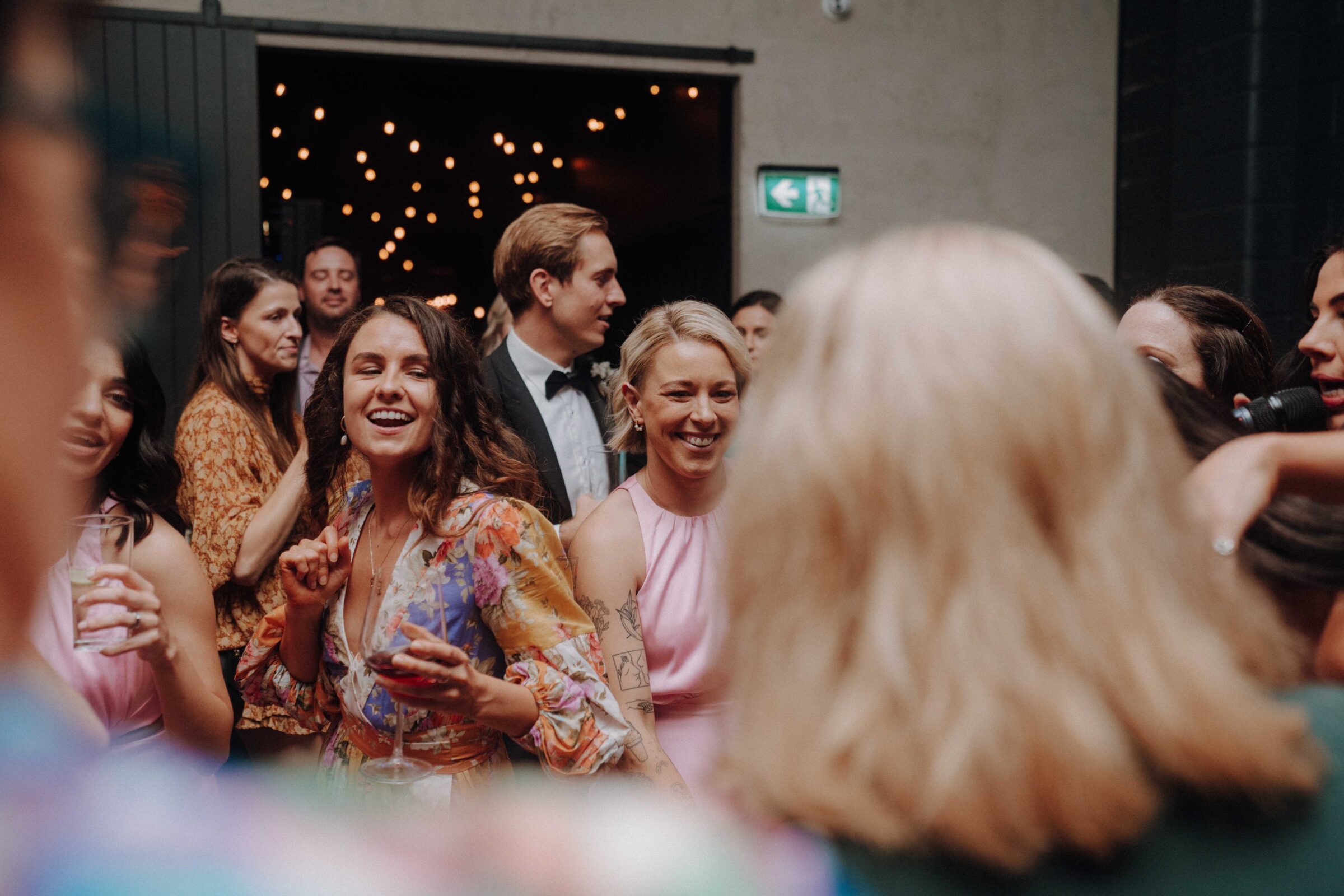 A group of people at an indoor event, some smiling and holding drinks. String lights are visible in the background.