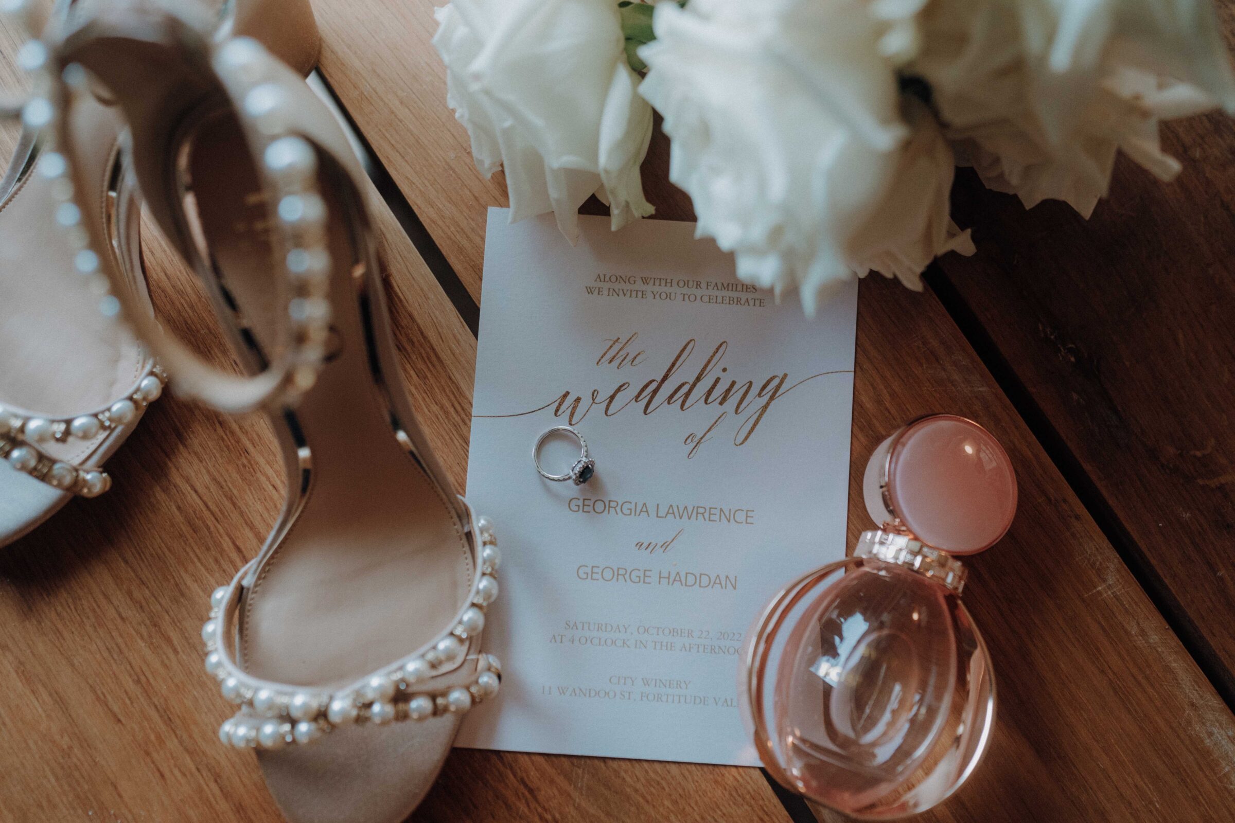 Wedding invitation with rings on top, surrounded by pearl-embellished heels, a perfume bottle, and white roses on a wooden surface.