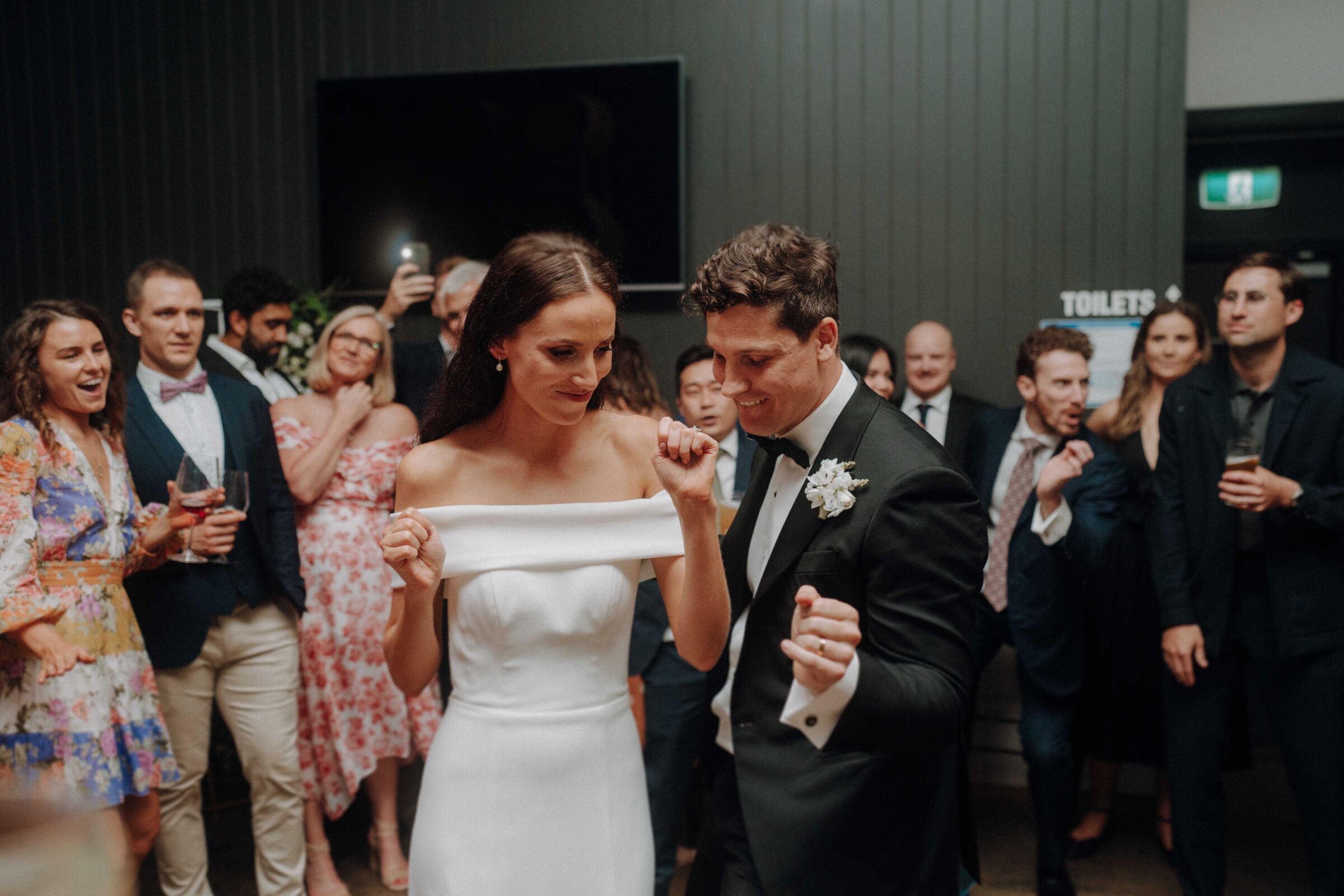 A bride and groom dance together at a wedding reception, surrounded by guests watching and taking photos.