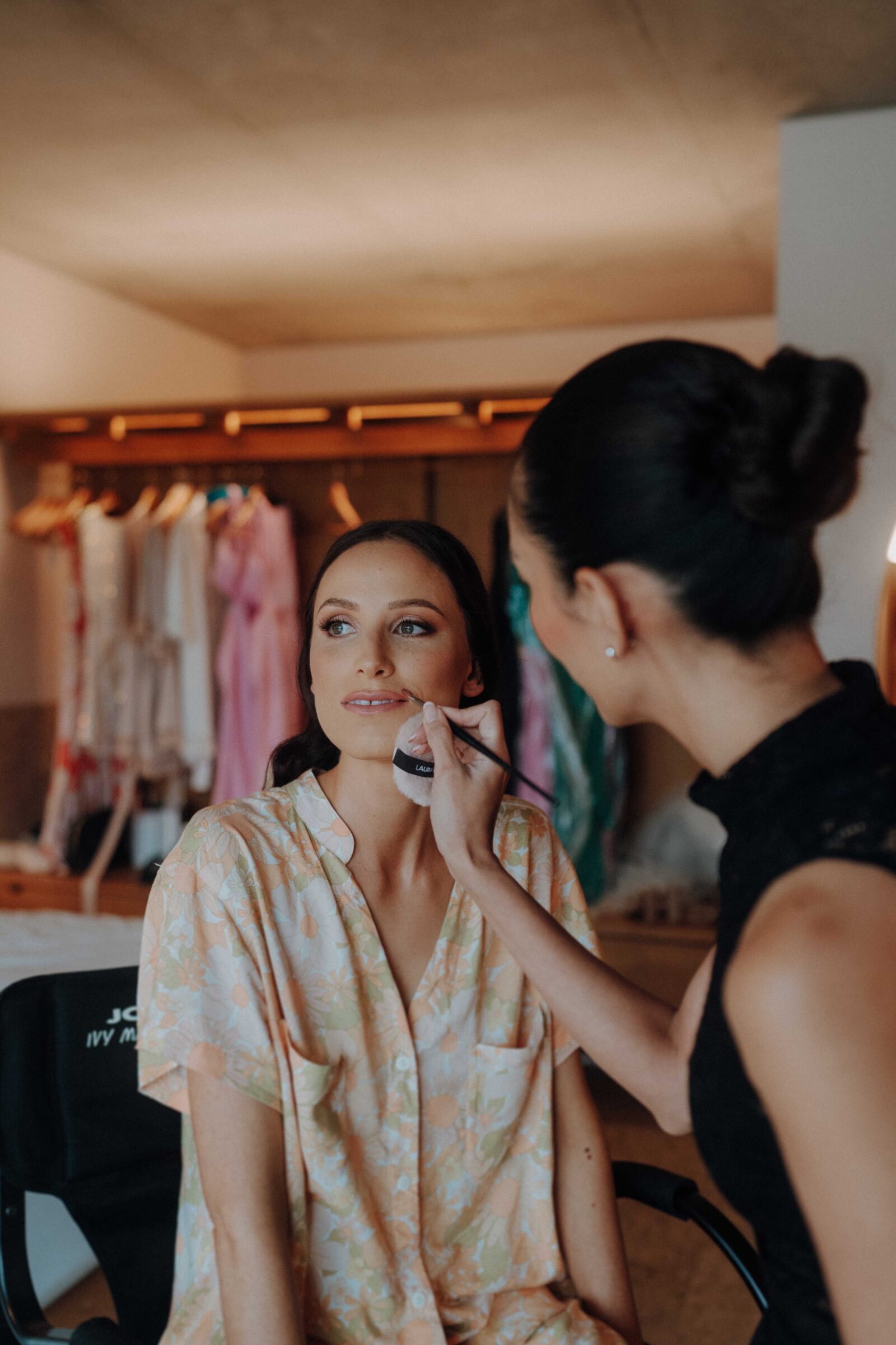 A woman in a patterned shirt sits while another woman applies makeup to her face in a dressing room with clothing hanging in the background.