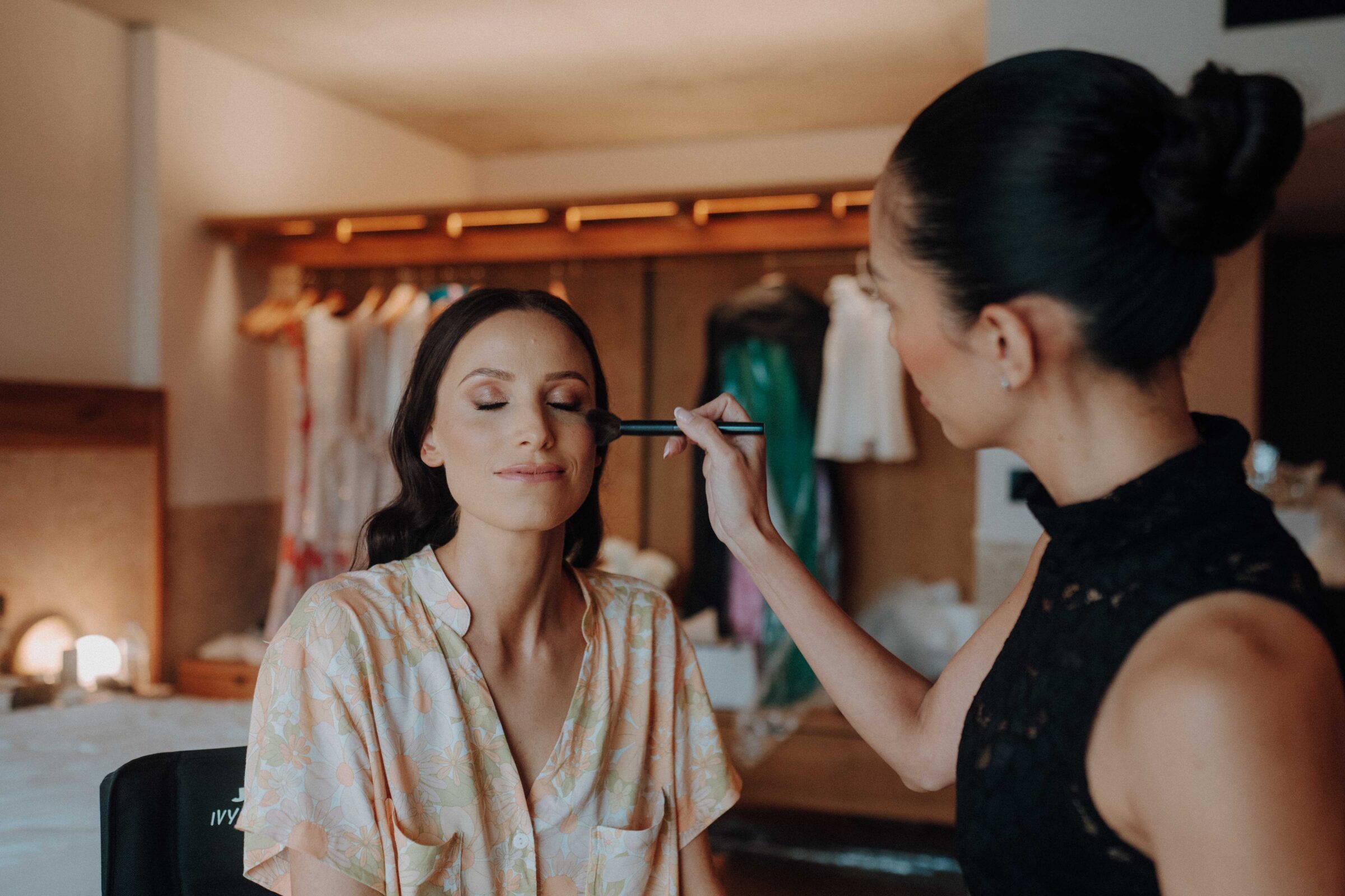 A woman in floral robe sits with eyes closed as another person applies makeup with a brush in a softly lit room.