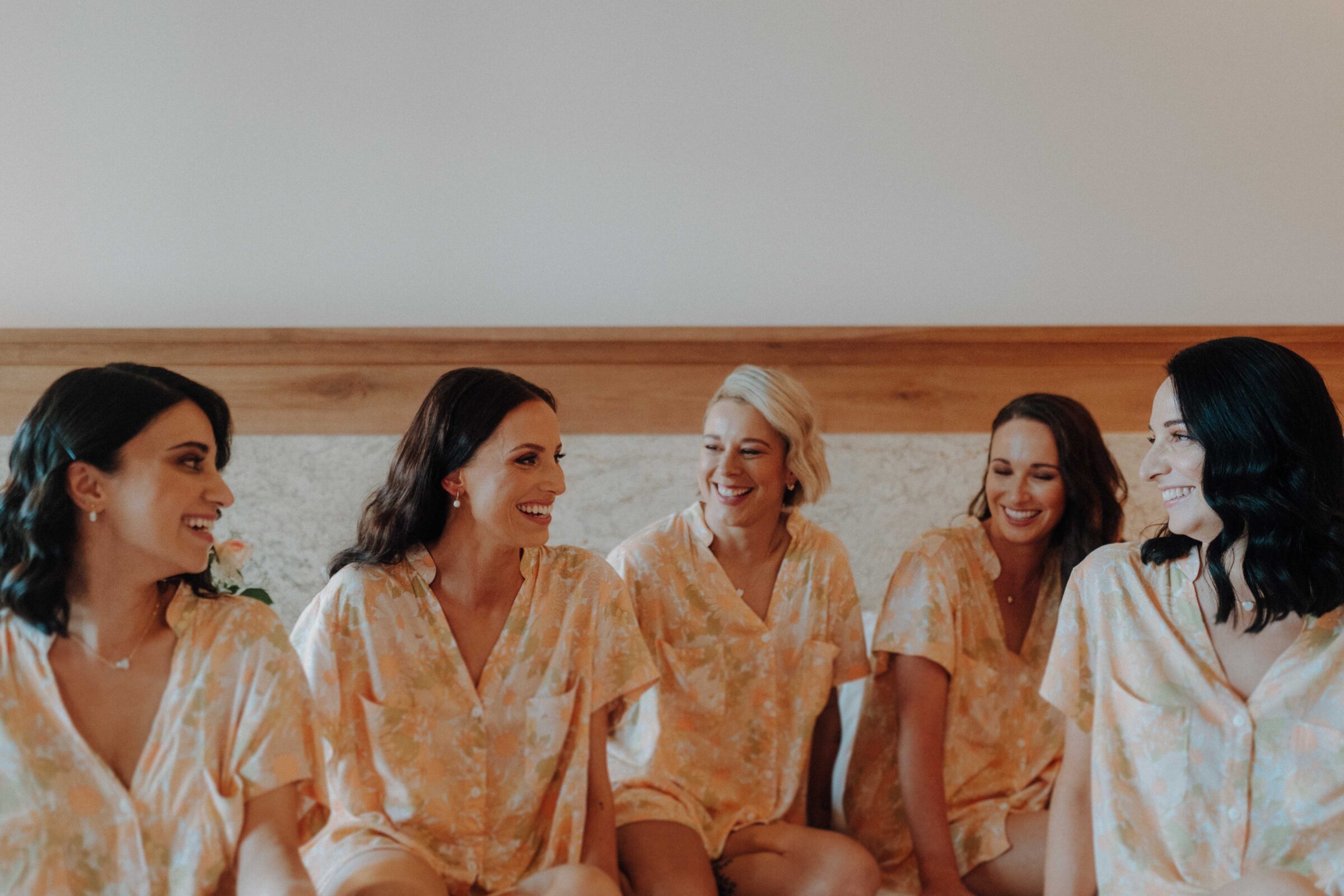 Five women in matching floral outfits sit and smile while chatting indoors.