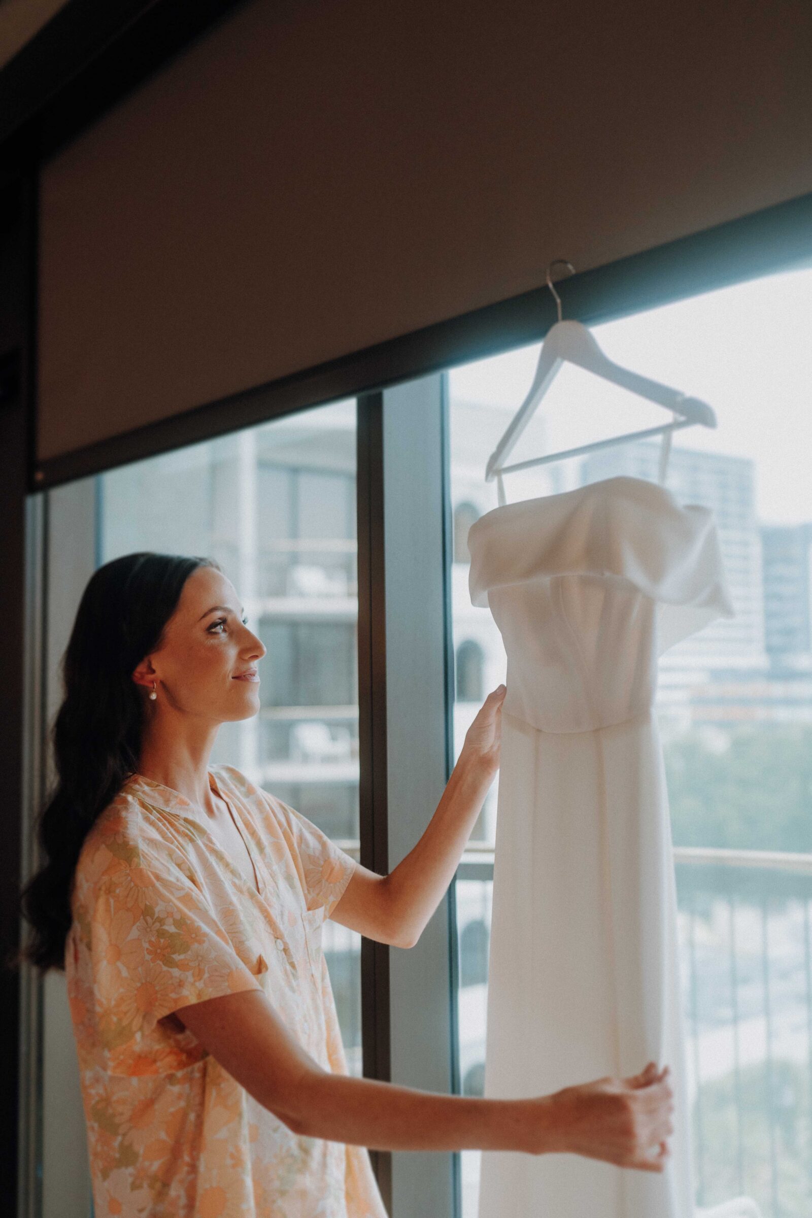 A woman stands by a window, observing a white off-the-shoulder dress on a hanger.