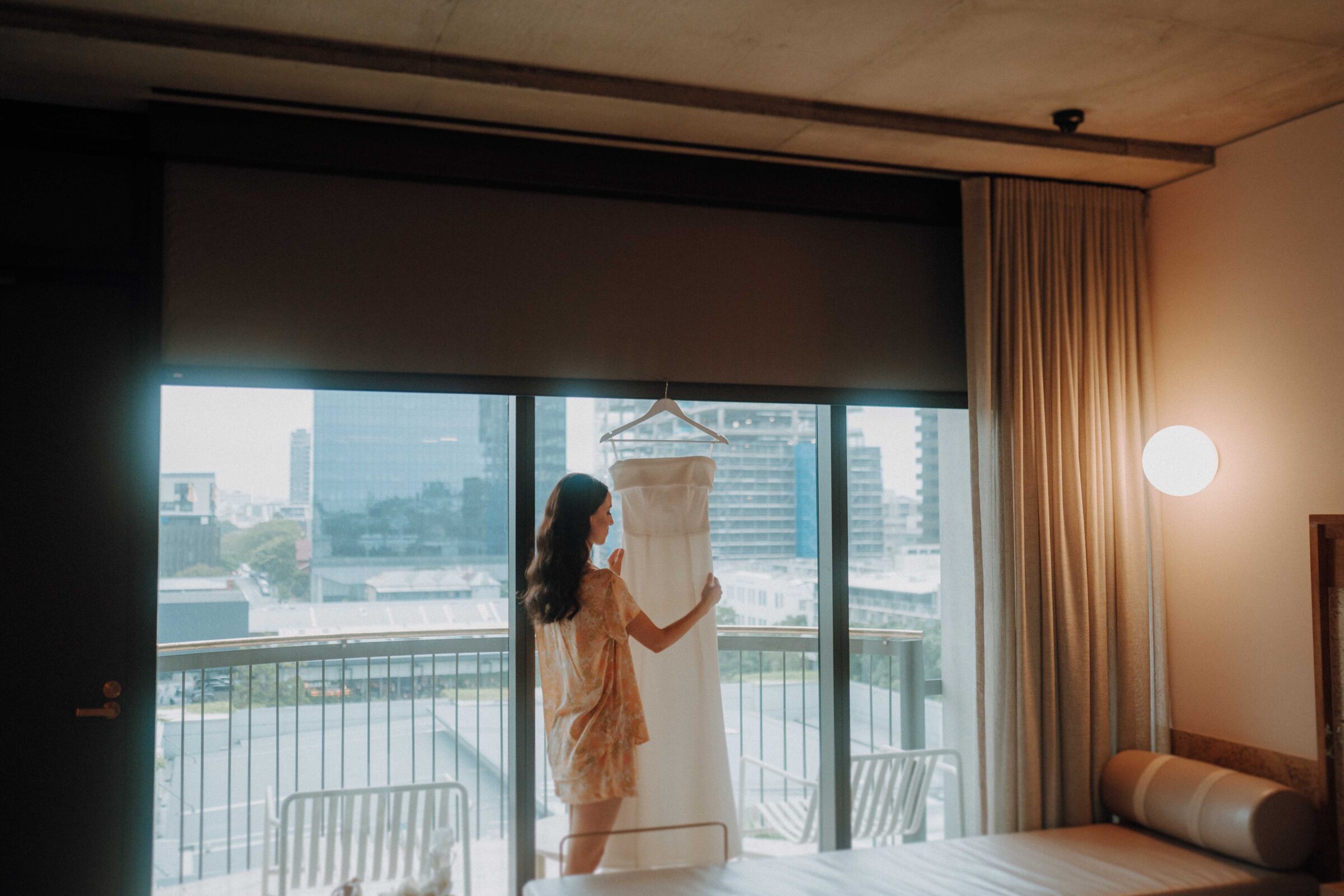 A woman stands in a room near a large window, holding a white dress on a hanger. The cityscape is visible outside.