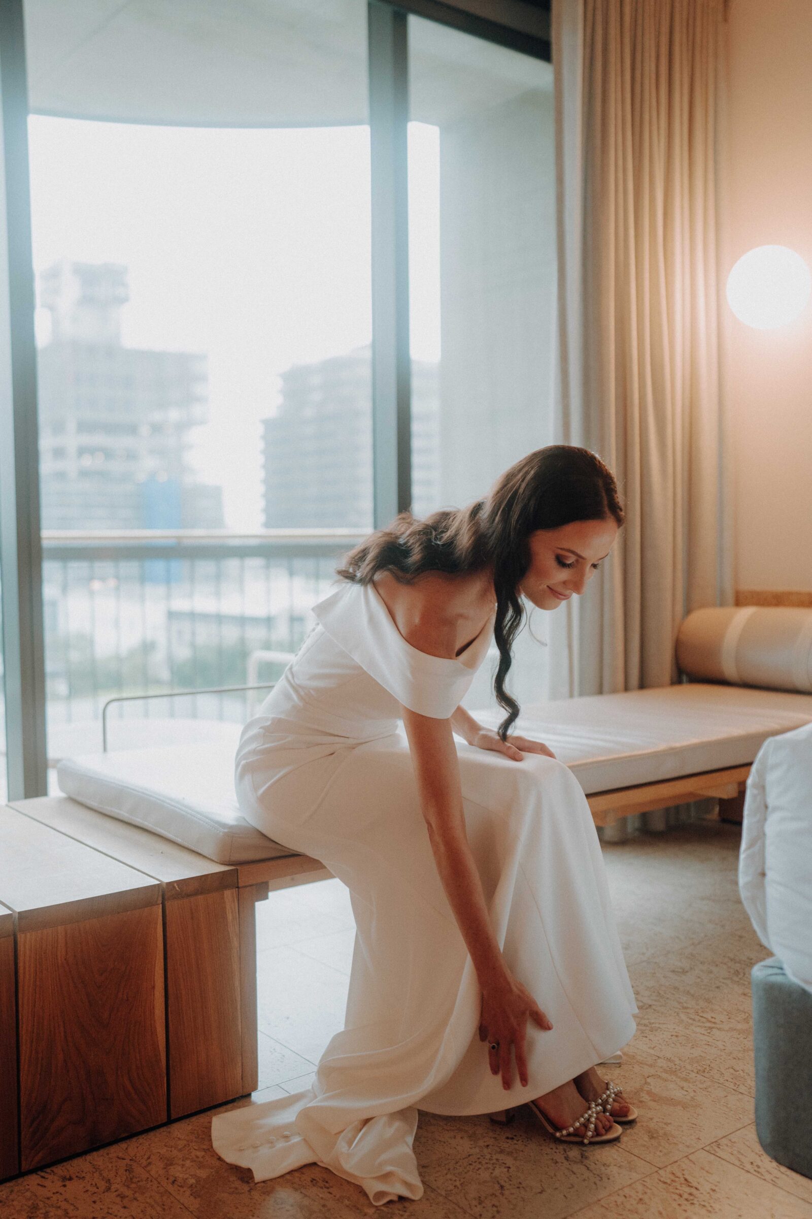 A woman in a white dress adjusts her shoe while sitting on a wooden bench in a modern room with large windows.