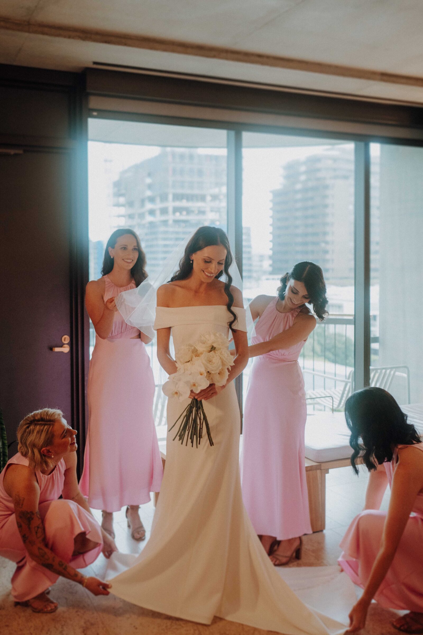 A bride in a white gown holding a bouquet stands with four bridesmaids in pink dresses adjusting her train indoors, with a city view in the background.