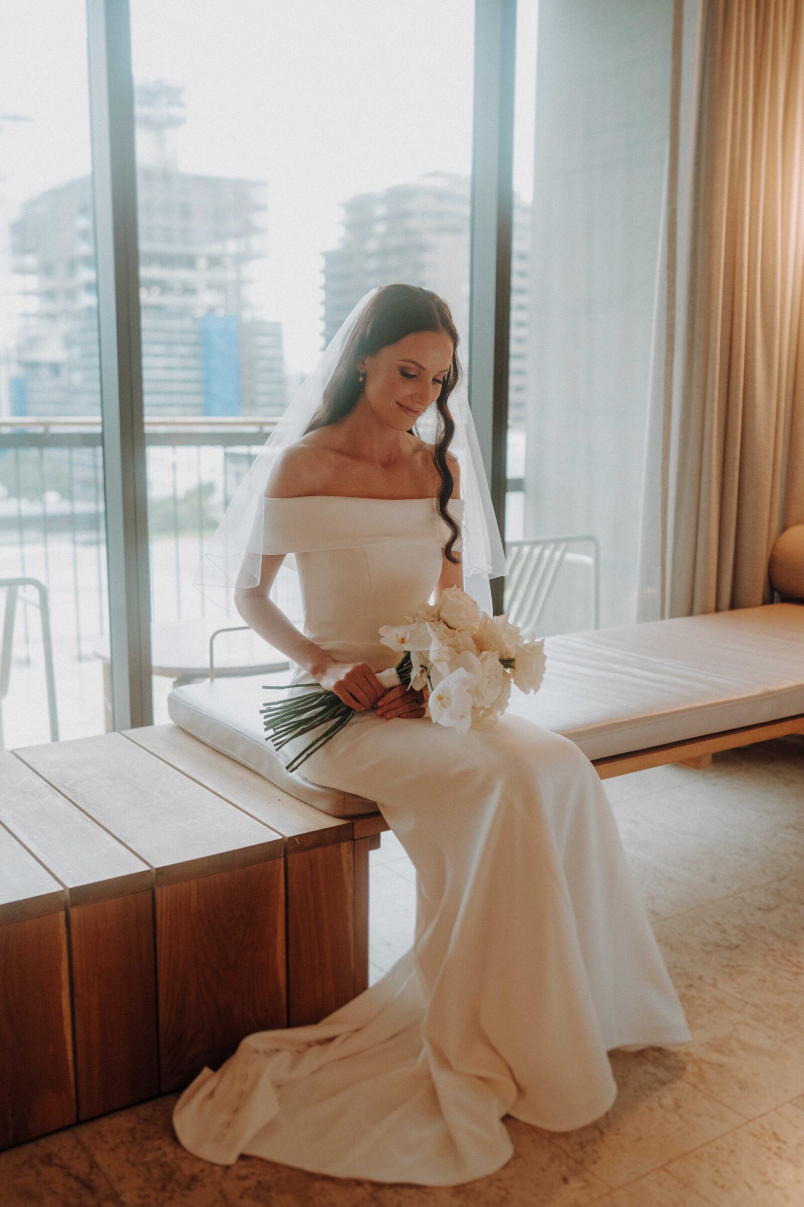 A bride in a white gown and veil sits on a bench indoors, holding a bouquet of white flowers, with large windows and city buildings in the background.