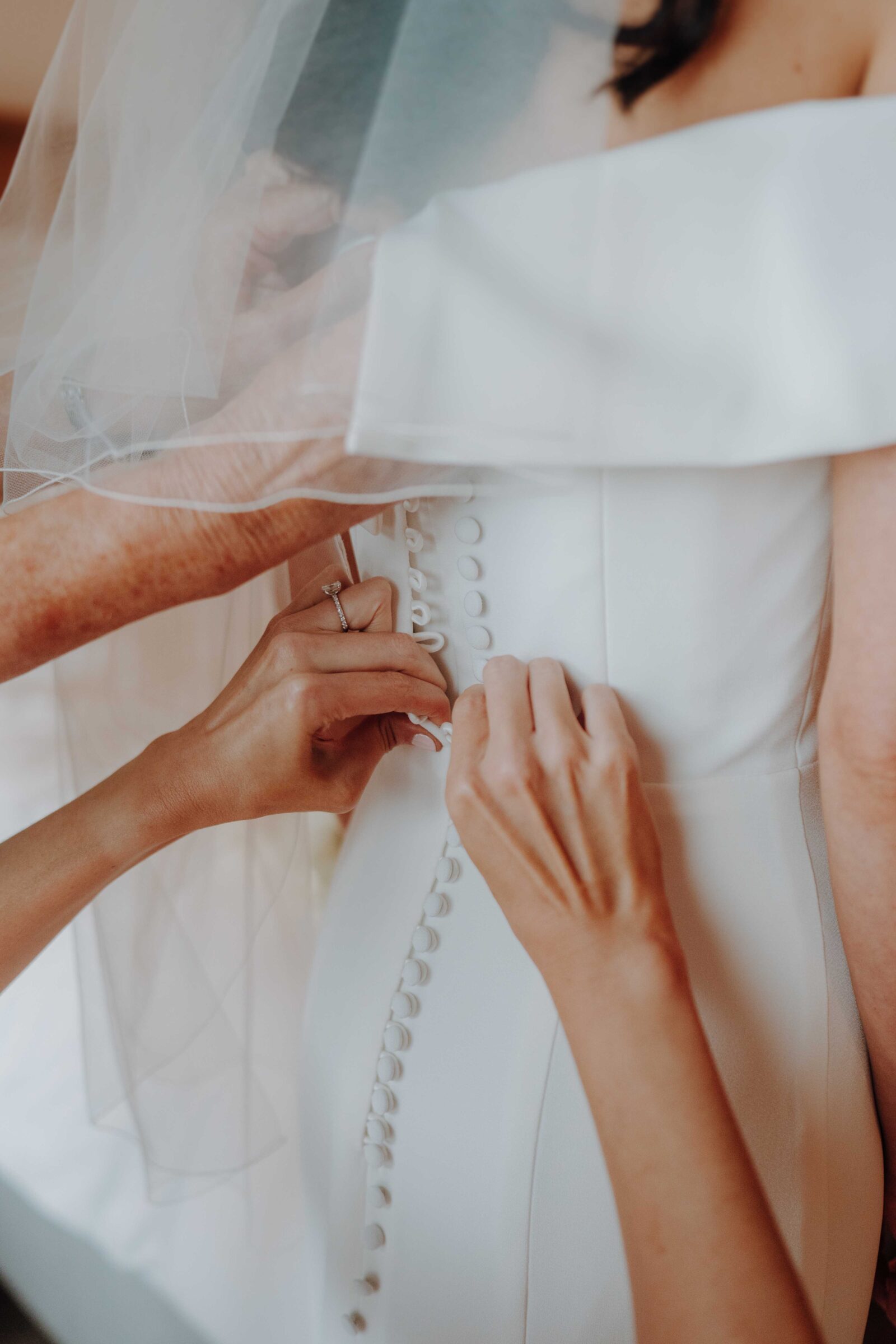 Hands fastening buttons on the back of a white wedding dress worn by a bride.