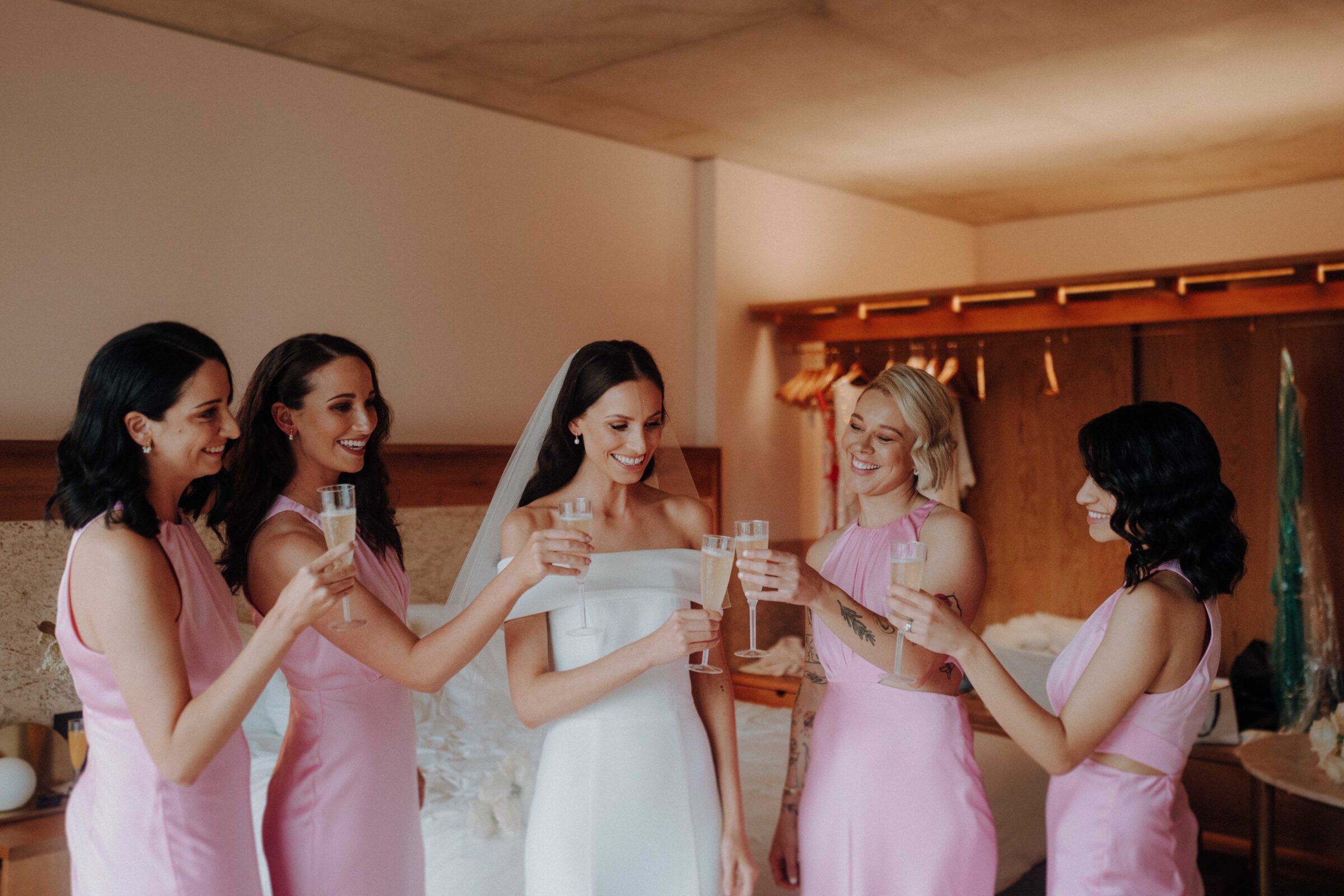 Five women in pink dresses toast with champagne flutes around a woman in a white dress in a room with wooden elements.