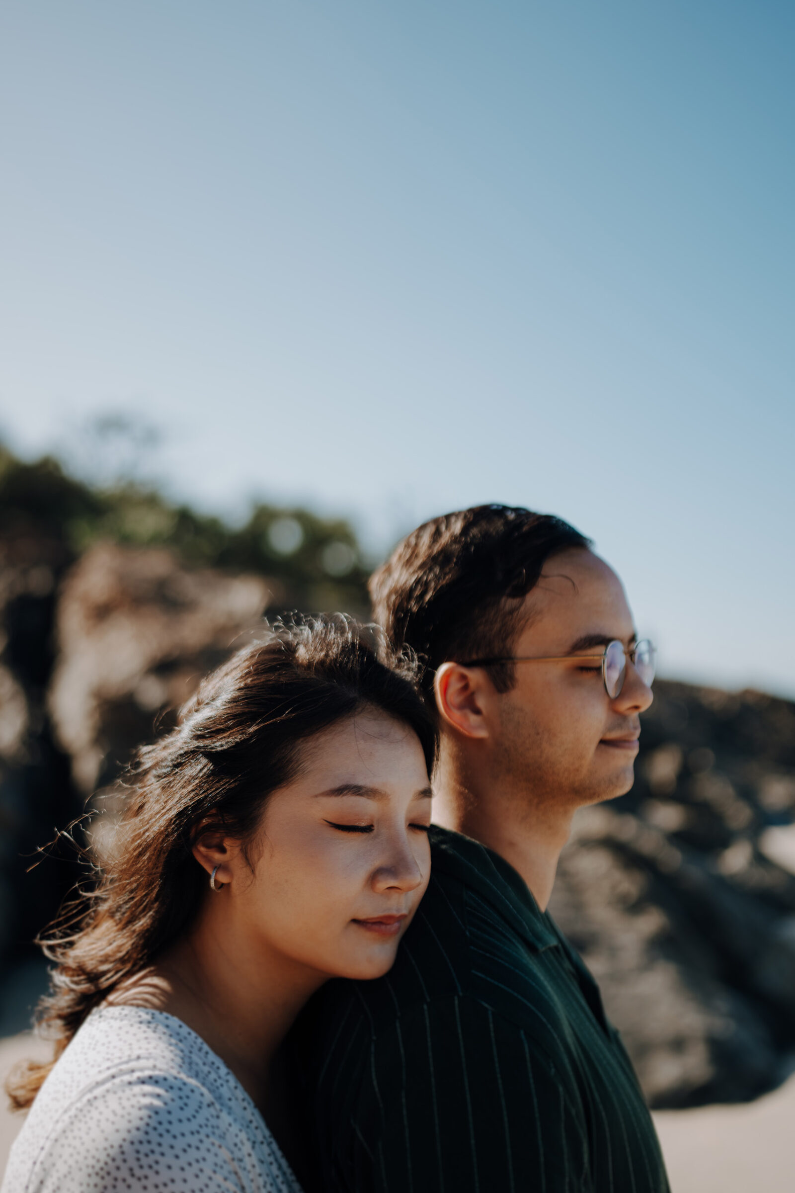 A woman leans against a man, both with closed eyes, standing outdoors against a rocky background under a clear blue sky.