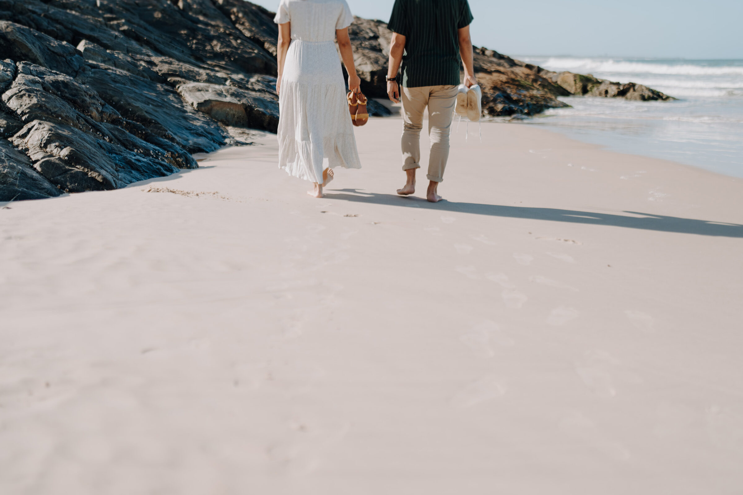 Two people walk barefoot on a sandy beach near rocks, holding sandals and a hat.